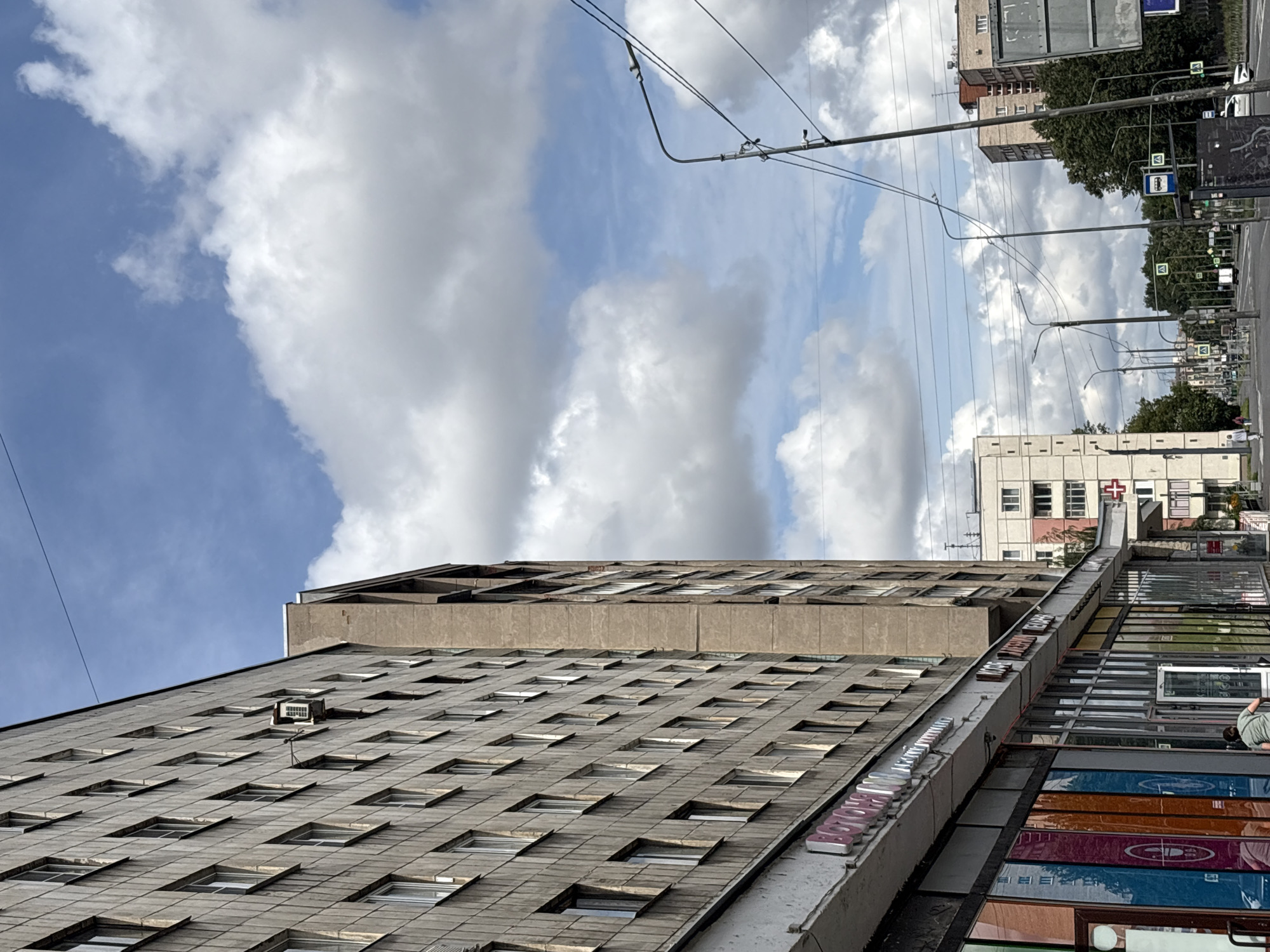 The image captures a street scene in Saint Petersburg, Russia, focusing on the exterior of a large, multi-story residential building with a concrete facade. The building's facade is adorned with many uniform windows arranged in a grid pattern. A single air conditioning unit is mounted on an external bracket on the building's wall. Below the residential units, a row of storefronts with large glass windows is visible. These shops display colorful signage and displays. A person is seen partially entering one of the shops. The street is lined with lampposts and electrical wires crisscross the sky, which is partly cloudy with a blue sky peeking through. In the background, other buildings and trees can be observed, suggesting an urban environment. A street sign with bus route information is also present.