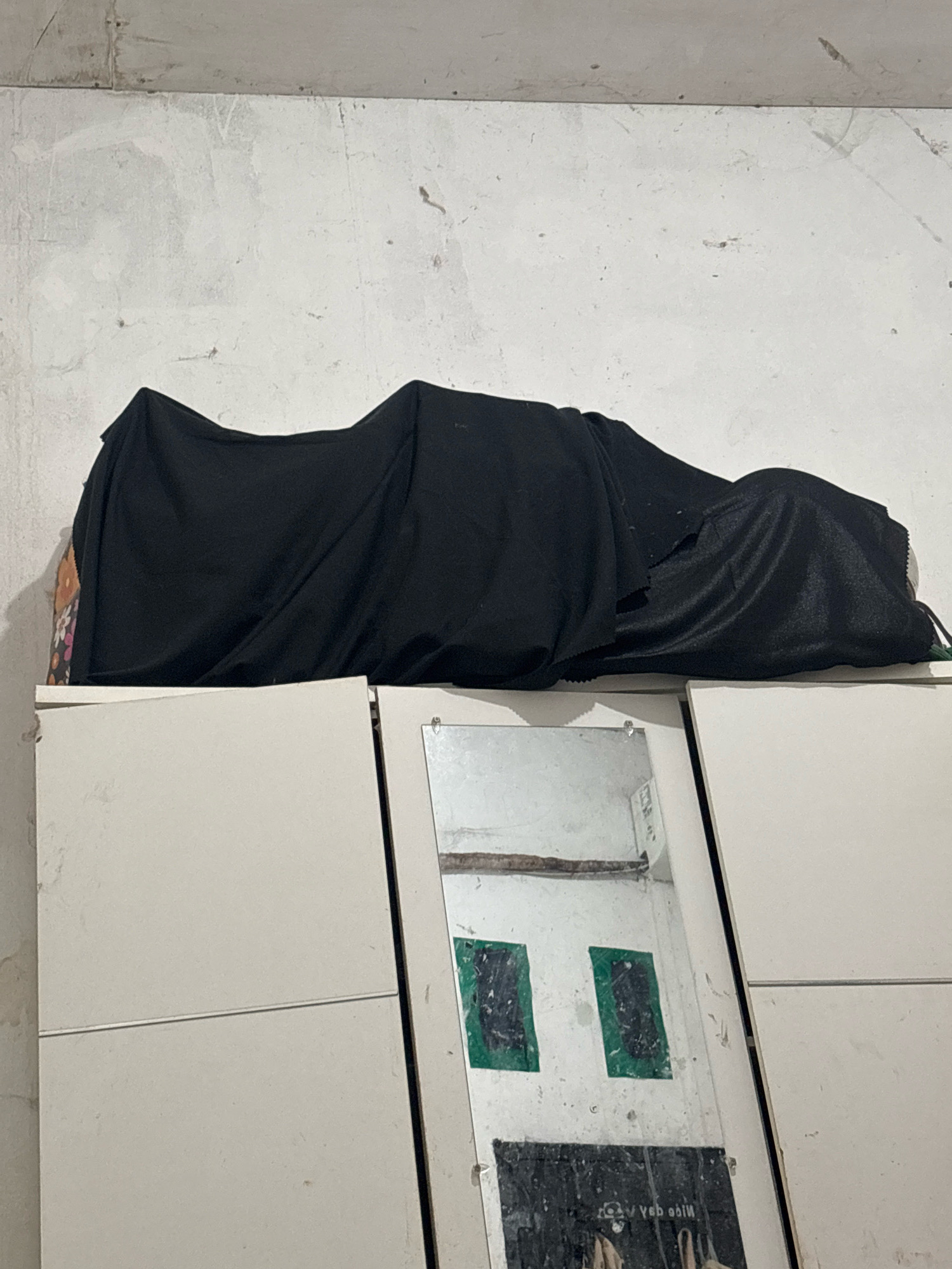 A black cloth covers an object that is lying horizontally on top of a white cabinet. The object appears to be an elongated shape, possibly a person or a large item. The cloth is draped loosely, with folds and wrinkles creating shadows. The cabinet has a plain, matte white finish and a horizontal seam across its front doors. A mirror is mounted on the front of the cabinet, reflecting a white wall with some debris. Two green rectangular shapes are visible in the reflection, as well as a poster with an upside-down reflection of text. The background wall is textured and appears to be made of concrete or plaster, with some marks and stains. This scene is located in North Jakarta, Indonesia.