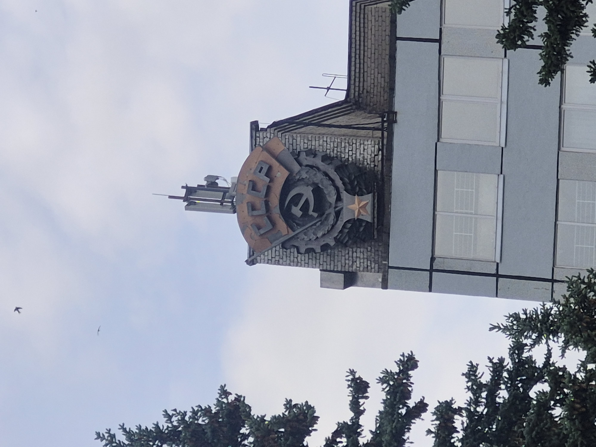 Here's a description of the image:
A low-angle shot depicts a building with a Soviet Union emblem prominently displayed on the top corner. The emblem is circular with the Cyrillic letters "CCCP" above a hammer and sickle. The emblem is mounted on a brick structure that juts out from the main building. The building itself appears to be a modern structure with gray paneling and rectangular windows. In the foreground, tree branches with green foliage partially obscure the lower portion of the image. Two birds are visible in the sky to the left of the building. The sky is overcast with light gray clouds.
The building is likely located in the city of Chaikovsky, Russia, based on the emblem.