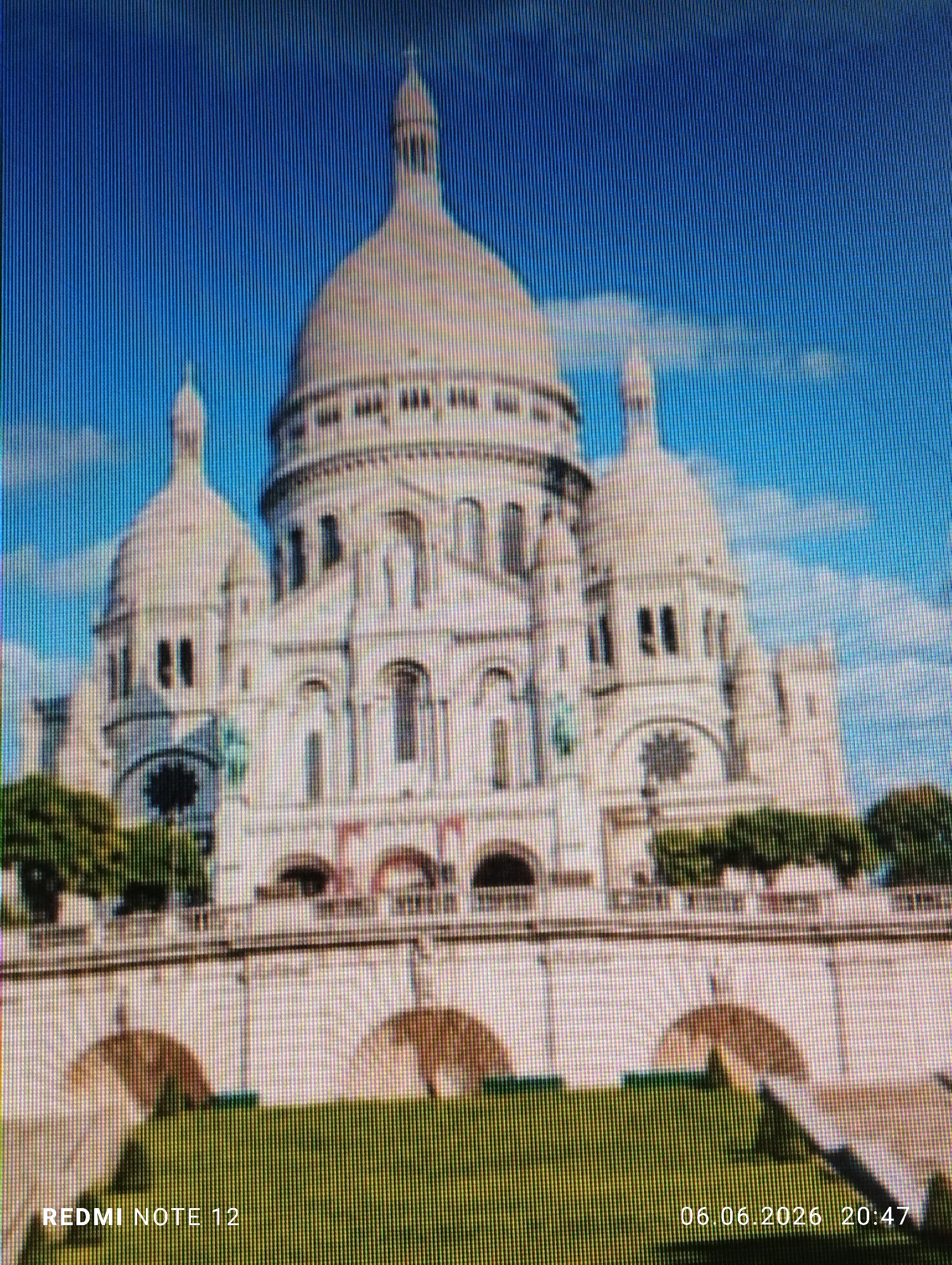 The image captures a large, ornate basilica, predominantly white, featuring a prominent central dome surmounted by a spired lantern, flanked by two smaller domes. The structure exhibits numerous arched windows, decorative details, and a multi-tiered design, crafted from light-colored stone. In the mid-ground, a wide, terraced stone platform with multiple supporting arches extends horizontally across the frame. Below this, a bright green, finely-manicured lawn occupies the foreground. The sky is a clear blue with scattered white clouds. No individuals are discernible within the scene. The scene is located in the city of Burgas, Bulgaria. The image itself displays artifacts consistent with a photograph taken of a digital screen, including distinct vertical banding and pixelation. Timestamp '06.06.2026 20:47' and device 'REDMI NOTE 12' are visibly watermarked in the lower right and lower left corners, respectively.