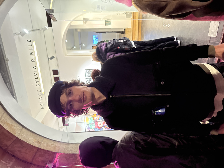 The image captures an indoor scene, rotated 90 degrees clockwise, depicting a person standing in what appears to be an art gallery or exhibition space. The primary subject is a person with wavy, dark brown hair and fair skin, wearing a dark beret and a dark (likely black) jacket with a shearling or fleece-lined collar. They are looking directly at the camera with a neutral, perhaps slightly observant expression. A small, dark blue rectangular object, possibly a security tag or a small camera, with white text is clipped to the left side of their jacket.
In the background, behind and to the left of the main subject, a large glass panel reflects the interior of the space. Printed vertically on this glass panel are the words "ESPACE SYLVIA RIELLE," indicating the name of the venue. Beyond the main subject, an arched doorway leads to another room or section of the gallery. Through this archway, several other people are visible, some with their backs turned or partially obscured. One person in the mid-ground wears a distinctive purple-patterned jacket. On the walls within the gallery, various artworks are displayed. Two dark, possibly metallic or sculptural pieces with intricate patterns are visible directly behind the main subject. Further back, through the archway, a vibrant, colorful artwork, possibly resembling stained glass or a mosaic with abstract or figurative elements (like a clown face), can be seen. Another distant artwork appears to emit a blue light.
The lighting is artificial, with spotlights visible on the ceiling, suggesting an indoor event, possibly during the evening or in an artificially lit daytime setting. Another person's head, wearing a dark hooded garment, is partially visible in the lower right foreground, suggesting others are also present near the photographer. The overall scene depicts a cultural gathering, likely an art exhibition or opening, where attendees are viewing art.