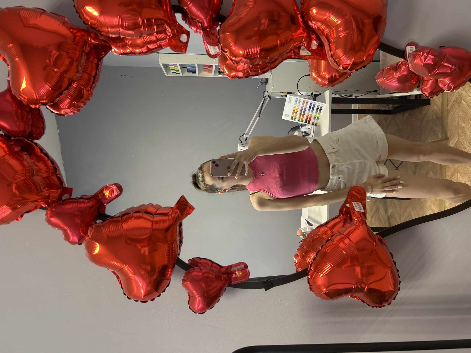 The image displays a young woman taking a selfie in a mirror that is prominently adorned with numerous shiny red heart-shaped foil balloons. The woman, reflected in the mirror, is wearing a pink sleeveless top and white shorts. Her face is obscured by the light-colored smartphone she holds up to capture the photo. She has light-colored nail polish on her fingers and toes and a small cross necklace. Her hair is tied back.
The red heart balloons are scattered around the mirror's frame and also visible within the reflection, suggesting they are part of the room's decor, creating a festive or romantic ambiance. Some balloons have small white tags with barcodes and partially visible text, including "P.I.T."
The background, as seen in the mirror's reflection, reveals an indoor setting with plain gray walls. A light-colored desk or table is visible, featuring a white swing-arm lamp and a color swatch chart with many small colored squares. Above the desk, shelves hold various neatly arranged items. The floor visible in the reflection is made of light-toned wooden planks. The lighting suggests a well-lit interior, possibly during the daytime or with bright artificial light.