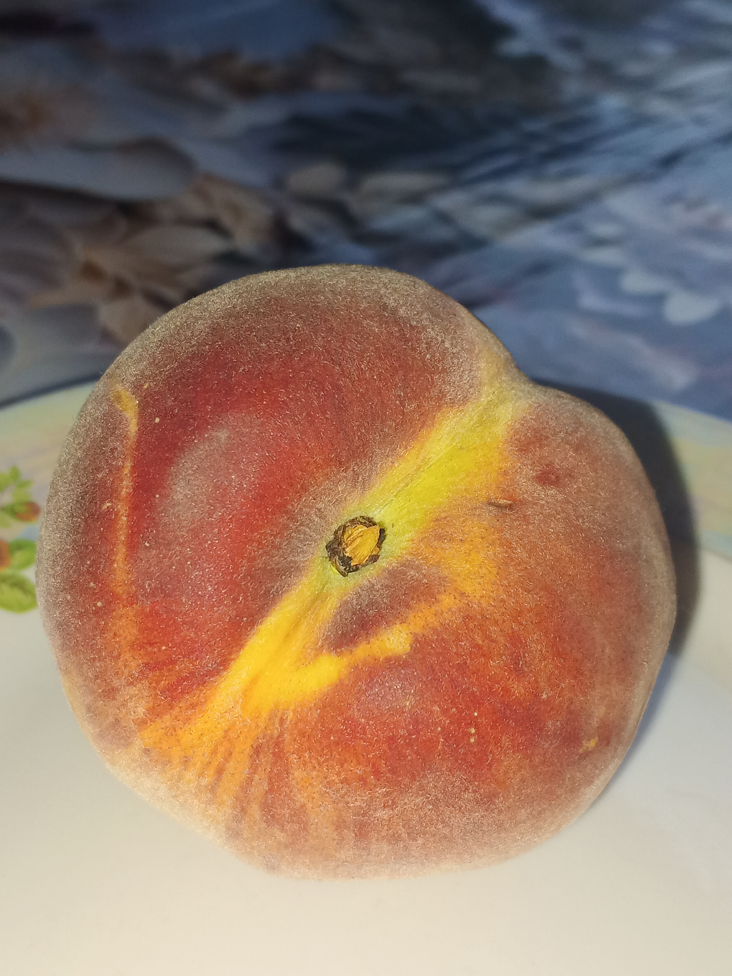A ripe peach is centered on a white plate. The peach has a fuzzy skin with patches of red and yellow. A shallow groove runs vertically down the center of the fruit, where the stem would have been. The background is blurred and appears to be a patterned fabric. There is no indication of the city of Sokhumi, Abkhazia, in this image.