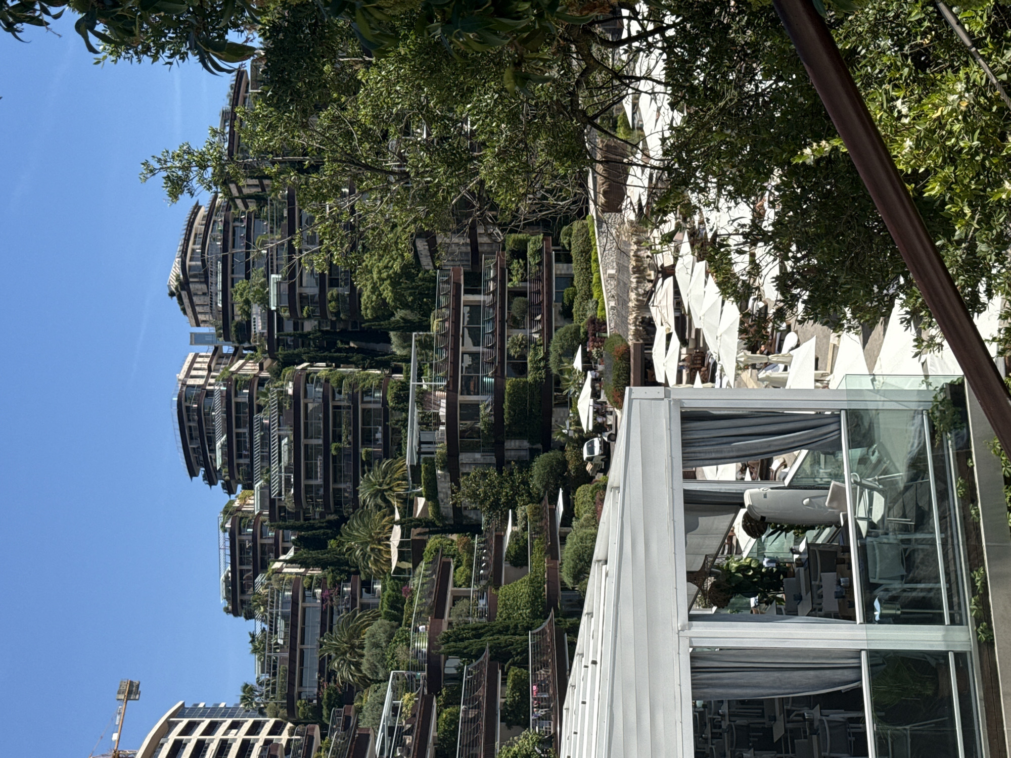 This is a high-angle, full shot taken on a bright, sunny day of a hillside residential complex with lush greenery in Budva, Montenegro. The architecture is modern and tiered, with multiple balconies and terraces covered in vegetation. The buildings are terraced down the hillside, with a distinct wave-like architectural style. In the foreground, a section of a white outdoor dining area is visible, with glass railings and tables and chairs set up inside. In the middle ground, there is a beach or pool area with numerous white umbrellas and lounge chairs. A small white vehicle, possibly a golf cart, is parked near an umbrella. The background is dominated by the sprawling residential complex and a clear blue sky. A construction crane is visible to the far left, suggesting ongoing development in the area. The scene conveys a sense of luxury and resort living.