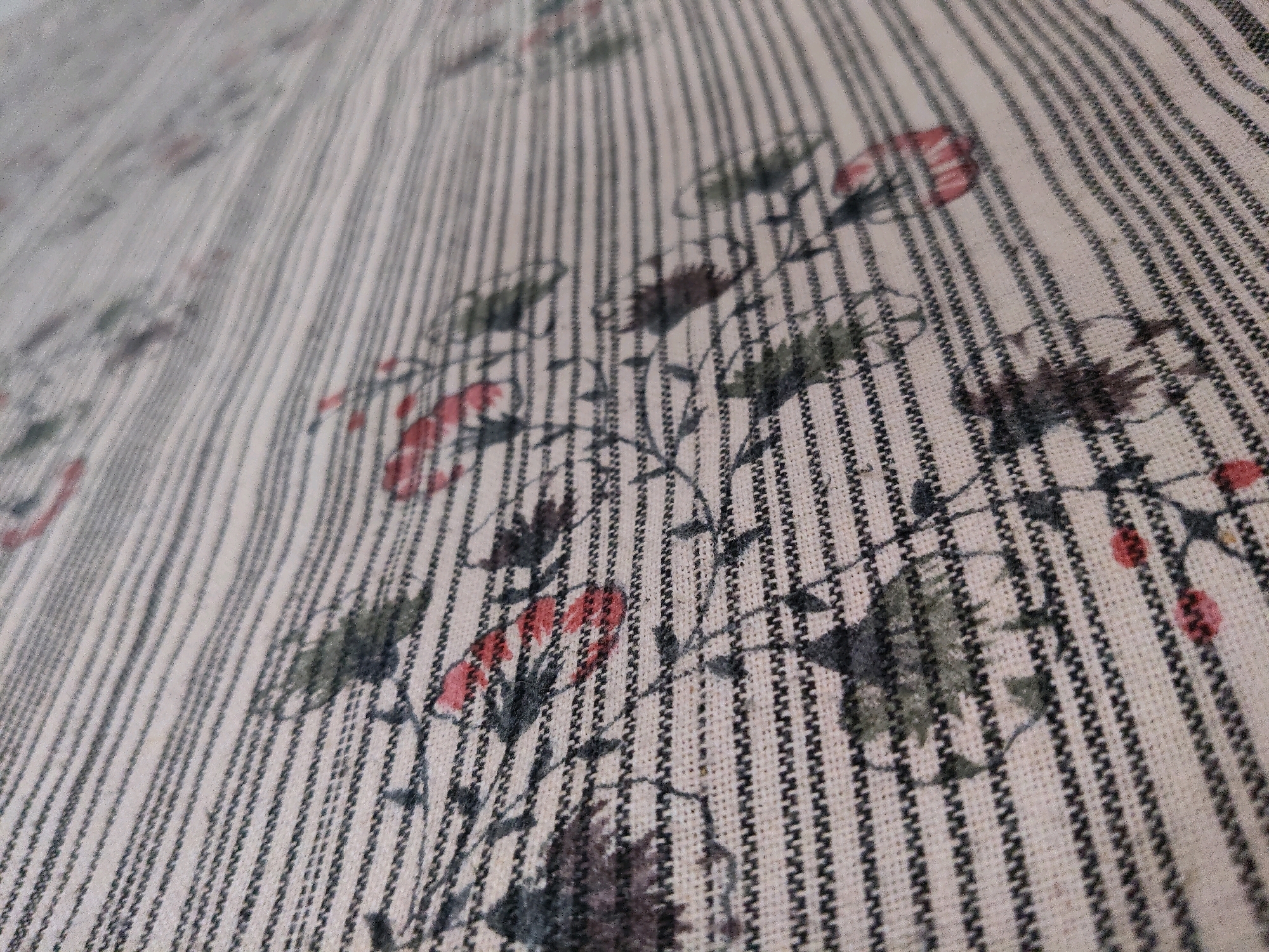 The image displays a close-up, angled view of a textured fabric with a subtle floral print. The fabric is predominantly a light cream or off-white color, with thin, dark vertical stripes running throughout. Overlaid on this striped pattern are delicate floral motifs in muted colors, including reds, greens, and dark purples. The flowers appear to be small and clustered, with some visible leaves and stems. The fabric appears to be a light to medium weight, and the texture is visible, suggesting it might be cotton or linen. There are no people or specific objects that would indicate a location within Moinabad, India; the image focuses solely on the textile itself.