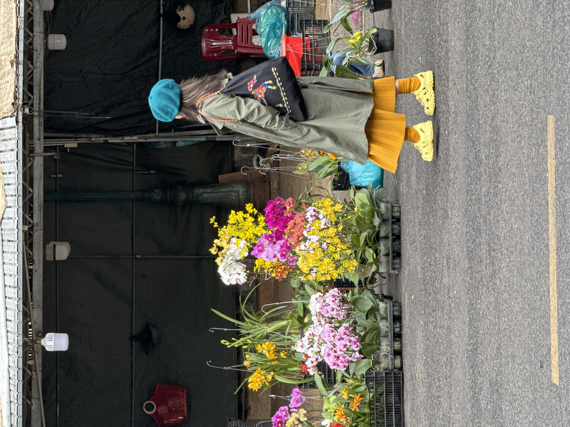 A person, viewed from behind and in profile, stands on an asphalt road in Da Lat, Vietnam, near a flower stall. The individual wears a teal beret, an olive green trench coat, a mustard yellow pleated skirt, yellow socks, and yellow clogs. They are carrying a black tote bag with a colorful embroidered bird design. The background features a stall with numerous potted orchids of various colors, including pink, white, yellow, and purple. The stall is under a corrugated metal roof, and behind it are dark green tarps, hanging lamps, and a red chair. A yellow line is painted on the asphalt road in the foreground. The scene appears to be an outdoor market.
