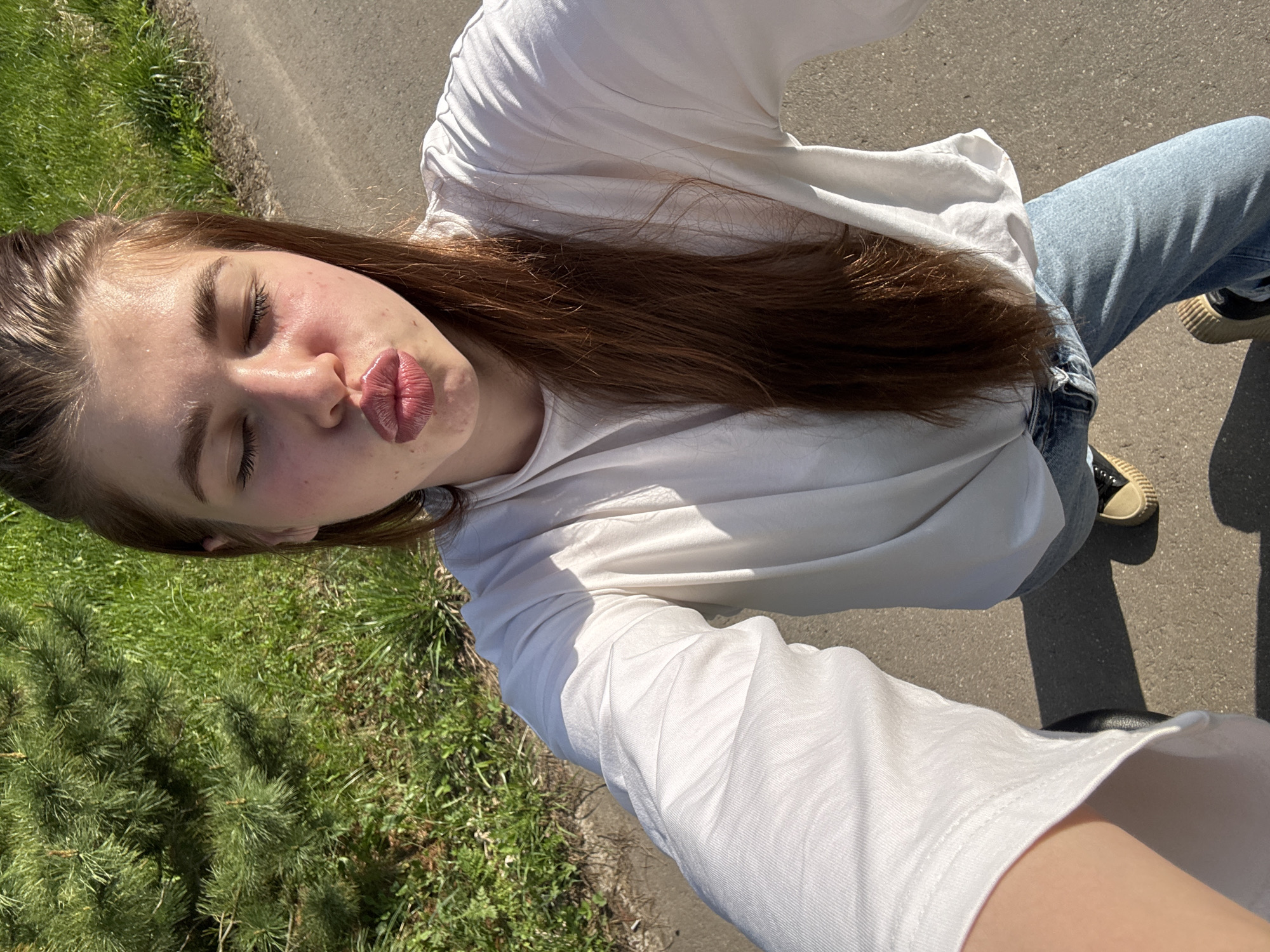 A young woman is taking a selfie outdoors on what appears to be a sunny day. She is wearing a white long-sleeved shirt and light blue jeans. Her hair is long and brown. Her eyes are closed, and she is puckering her lips as if blowing a kiss. The background includes a paved area and a grassy area with small pine trees or bushes. The exact location within Мещёры, Russia, cannot be determined from the image alone.