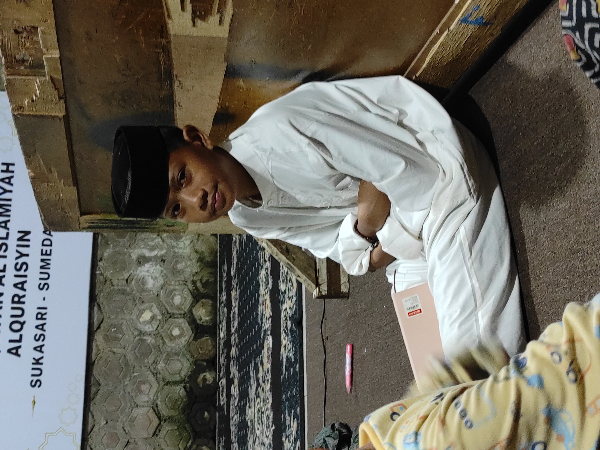 A young boy wearing a black peci cap and white attire sits with his arms crossed on the floor, leaning against a wooden structure. He is positioned indoors, as indicated by the textured wall and patterned carpet visible in the background. A sign behind him reads "ALQURAISYIN SUKASARI - SUMEDANG," suggesting the location is in Sukasari, Sumedang, Indonesia. In the foreground, a pink object, possibly a pen, lies on the floor, and another person in yellow pajamas is partially visible. The lighting is dim, creating shadows and highlighting the boy's face.