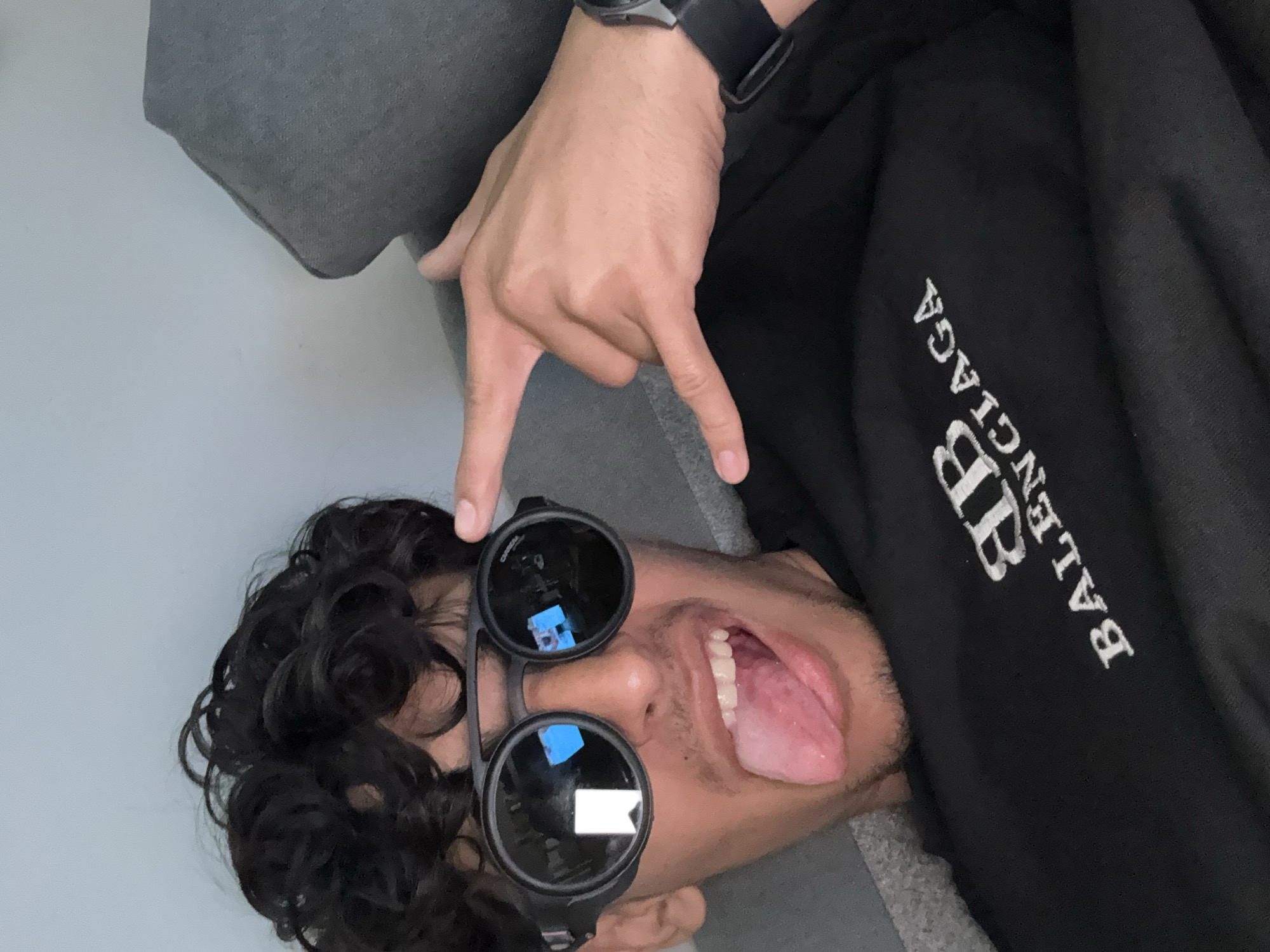 A person lies on a gray surface, possibly a couch or floor, wearing round, black sunglasses and a black Balenciaga hoodie. The person is sticking out their tongue and making a hand gesture. They are wearing a watch on their left wrist. The environment consists of a light gray wall. The location in the city of Kazichene, Bulgaria is not determinable.