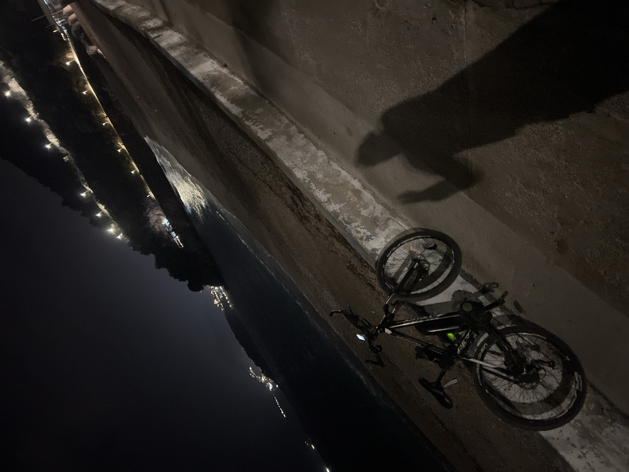 The scene is an angled, low-light shot taken in Boreti, Montenegro, at night. A bicycle lies on its side against a concrete wall. The wall runs diagonally across the upper half of the frame. A dark shadow, possibly of a person, is cast on the wall. In the background, across what appears to be a body of water, are indistinct lights indicating a town or city. The left side of the image is dark, showing what seems to be an embankment with sparse lighting.