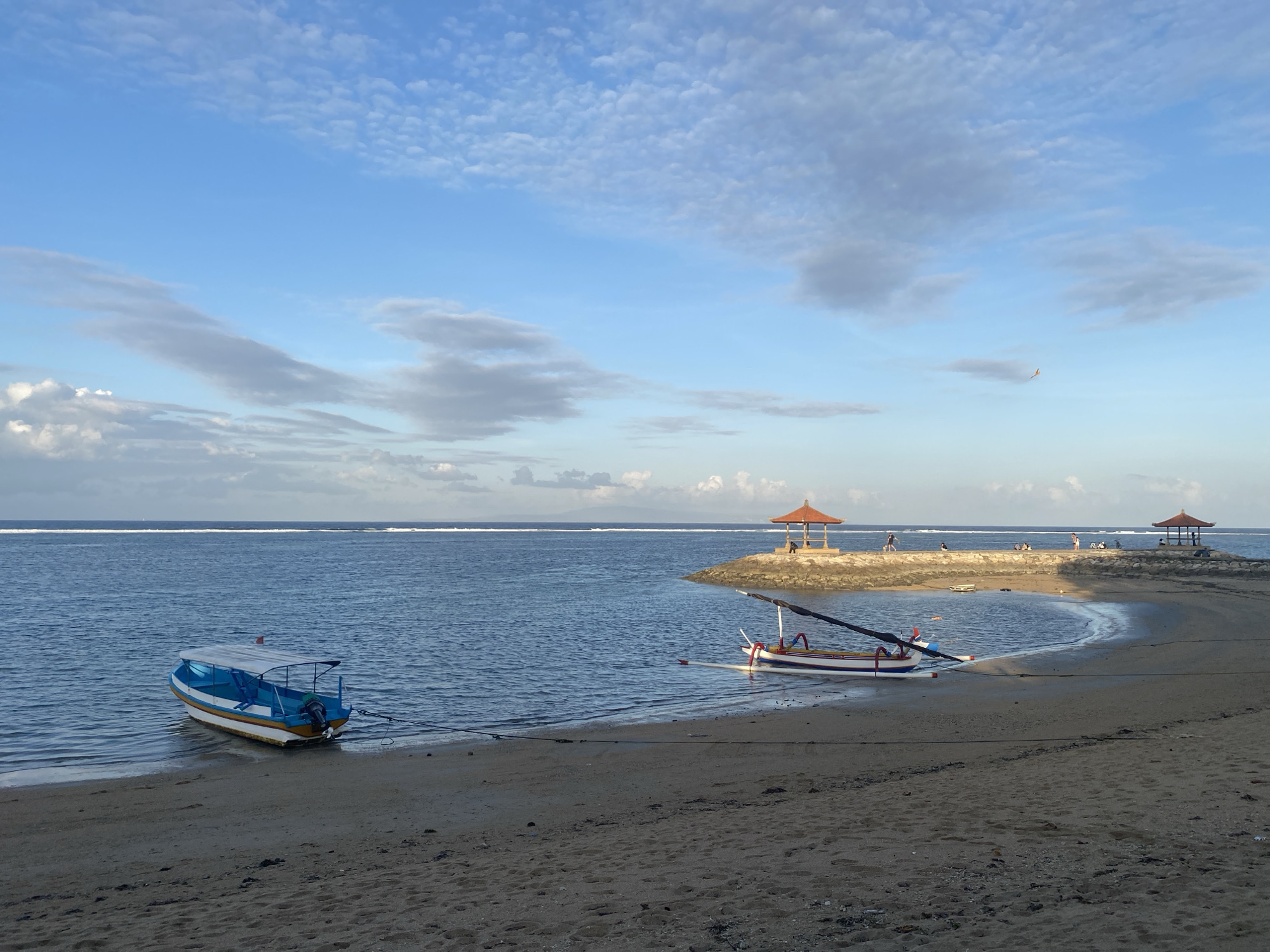 The image shows a beach scene in Denpasar, Indonesia, likely at sunrise or sunset given the soft lighting and the color of the sky. In the foreground, a sandy beach slopes down to the water. Two boats are visible. The first, a white and blue motorboat with a canopy, is partially in the water and tied to the shore. To the right, a traditional Balinese outrigger boat, painted white with red and blue accents, rests on the sand, also tied down.
The water is calm and a deep blue, with a thin white line of waves breaking in the distance, suggesting a reef or breakwater. Further along the coastline, a man-made structure juts out into the sea. It consists of a stone pier leading to two open-air pavilions with orange, conical roofs. Several people are visible on and around the pier, some standing, others sitting, and one or two walking. A small kite is visible flying in the upper right portion of the sky. The sky itself is a gradient of light blue, with scattered white and gray clouds, some wispy and others more defined. The overall impression is one of tranquility and a popular tourist spot.