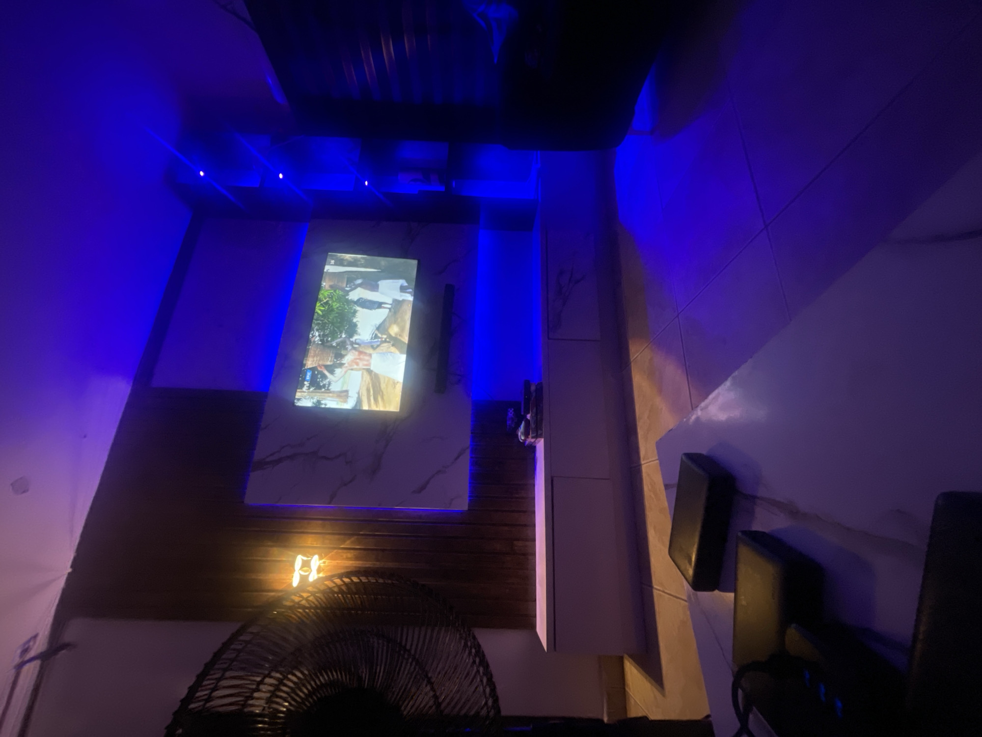 The image shows an interior space with blue ambient lighting. A flat-screen TV is mounted on a marble-patterned wall with wood paneling below. The TV displays an image with natural scenery. A black soundbar is mounted vertically to the right of the TV. A white cabinet is present to the right, with speakers placed on it. In the foreground, a black fan is partially visible. The image is angled, giving a distorted perspective. The location is unknown, but the city is said to be Lokoja, Nigeria.