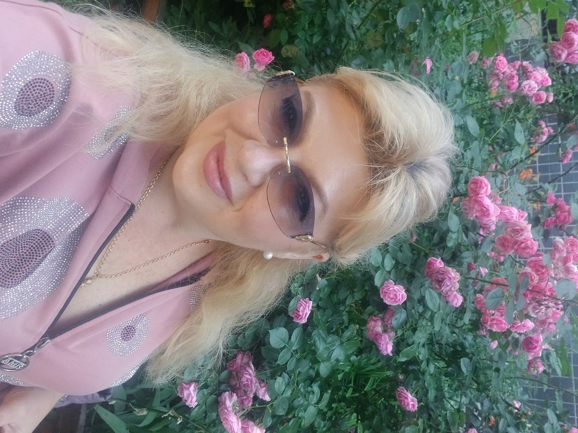 A middle-aged woman with blonde hair and fair skin, wearing sunglasses, a pink zip-up hoodie adorned with rhinestones, and a gold necklace, poses for a selfie in front of a bush of pink roses. The background appears to be a garden or park setting, with lush green leaves and numerous pink blossoms. The woman is smiling faintly. The overall scene is brightly lit, suggesting it was taken during the daytime. The photograph was taken in Odesa, Ukraine.