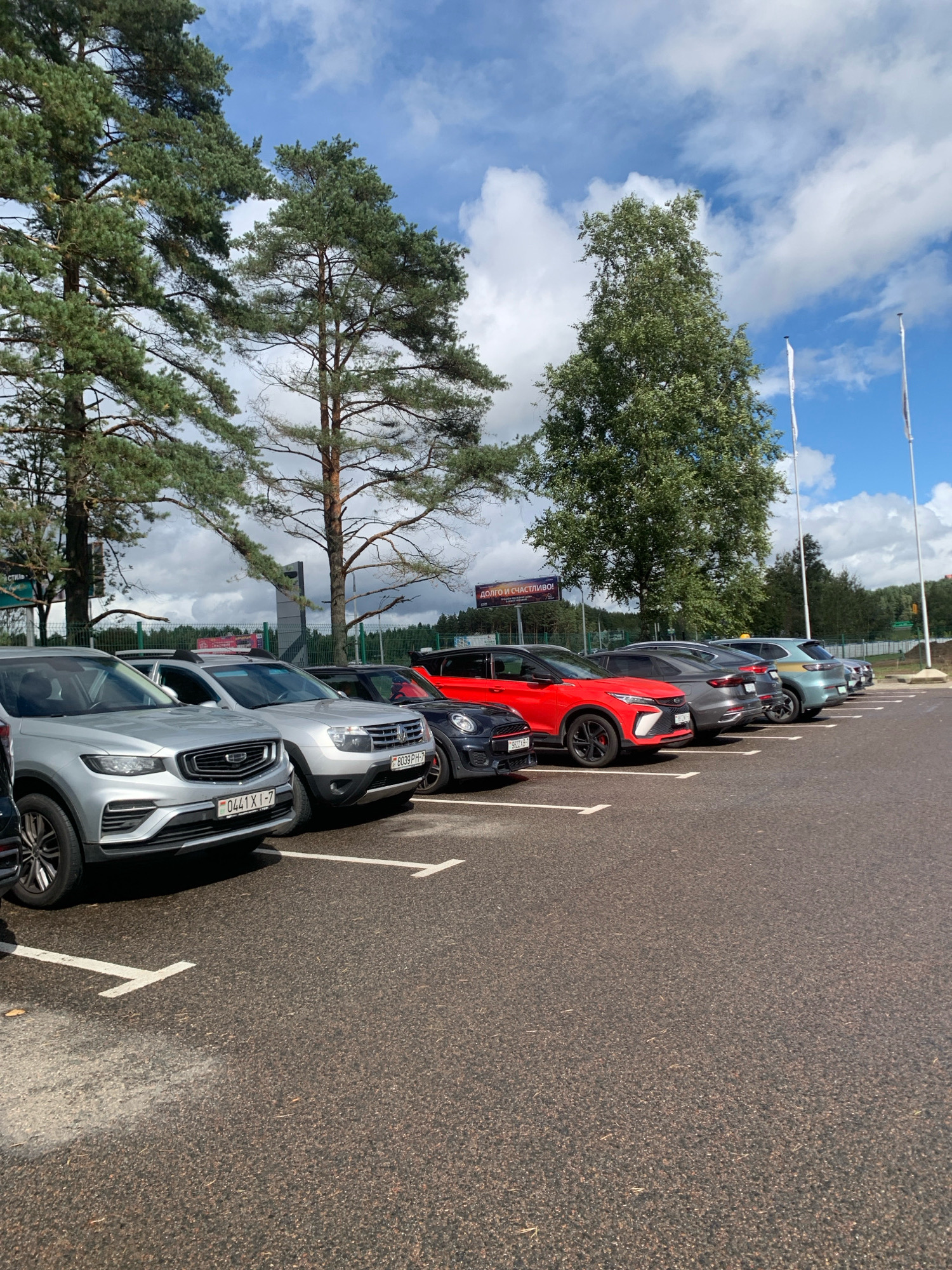 An outdoor, eye-level, full shot shows a row of parked cars on a paved lot under a partly cloudy sky. The cars are lined up nose-out in parking spaces marked with white lines. From left to right, the visible cars are a silver Geely SUV, a silver Dacia Duster, a dark colored hatchback, a red Geely SUV, a gray Geely SUV, a light blue Geely SUV, and a dark colored SUV. The license plates on the silver Geely SUV read "0441 X I-7". The license plate on the Dacia Duster reads "8039 PH-7". A large billboard with Russian text is visible in the background behind the cars. Two white flags are on poles to the right of the billboard. The lot is bordered by trees and a green fence. The pavement is dark and appears to be asphalt. The image is taken on a bright, daytime scene.