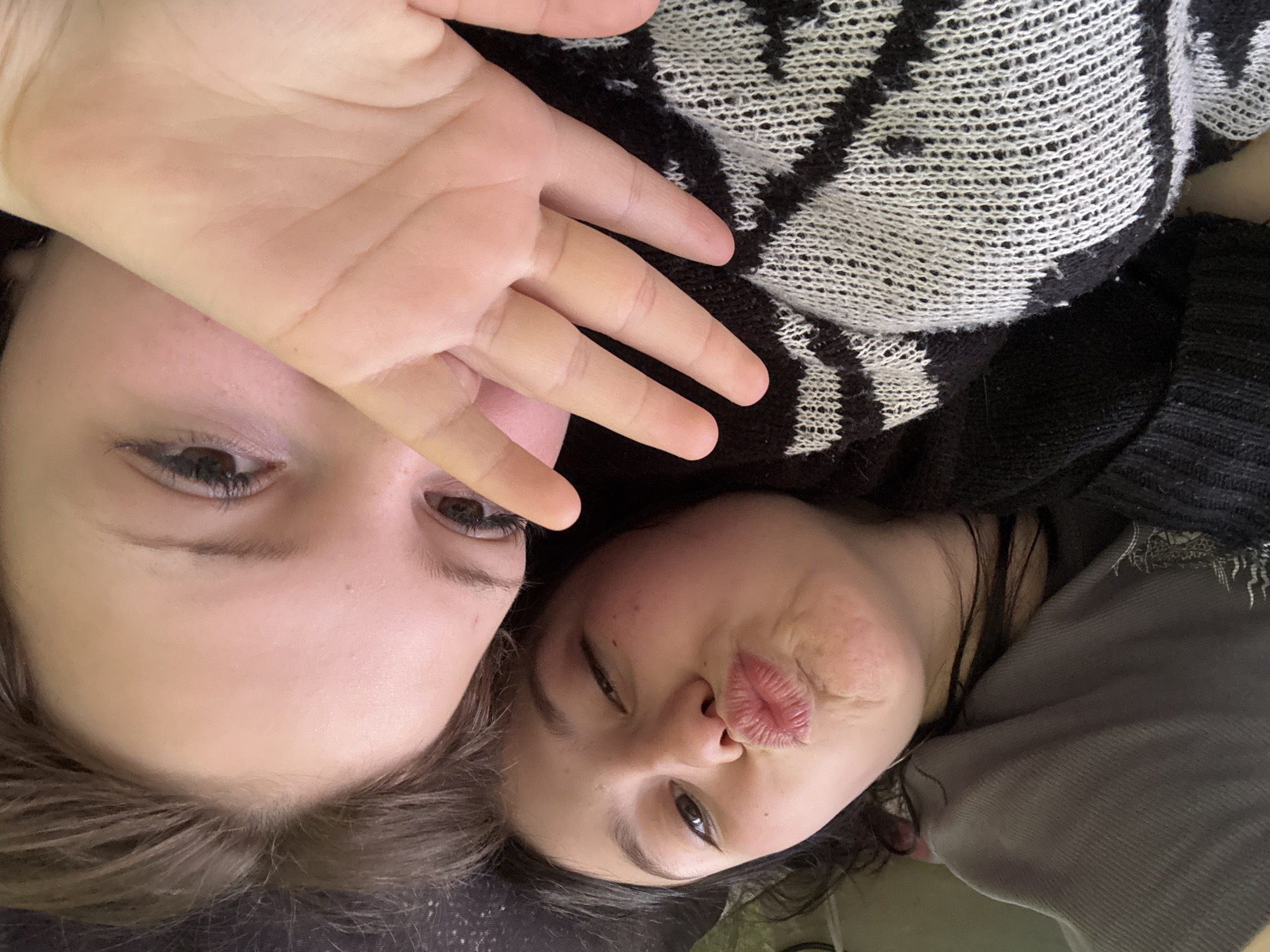 Two young women are lying down next to each other, posing for a selfie. The woman on the left has brown hair and is holding her hand up in front of her face. The woman on the right is making a kissing face. They are both wearing casual clothing. The background is blurry. There is no indication of Фоки, Russia in the image.