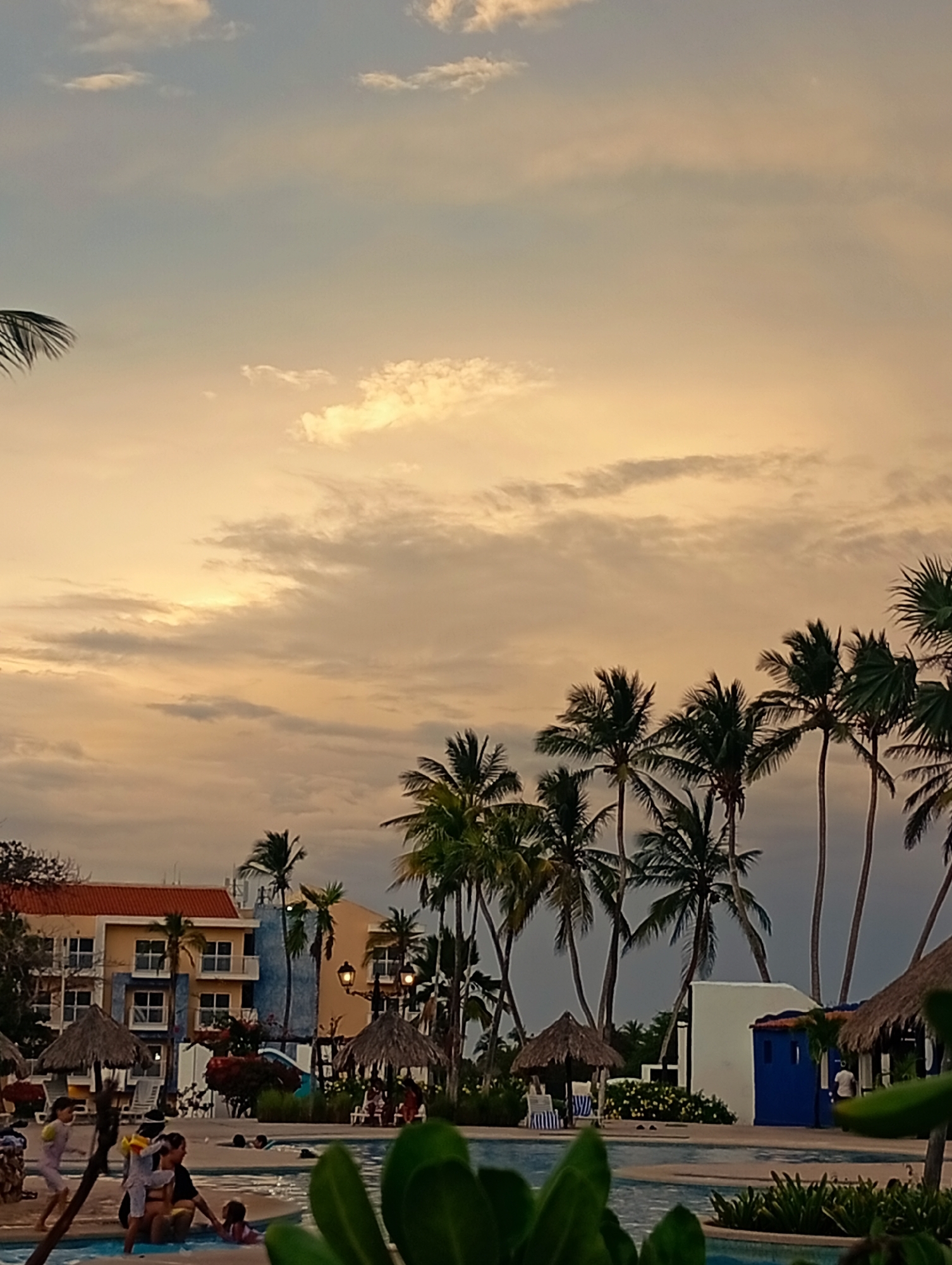 The image depicts a sunset scene at a resort in Playa El Agua, Venezuela. In the foreground, a swimming pool is visible with people, including adults and children, swimming and interacting near the edge. To the left, several individuals are seated or standing near the pool's edge.
Behind the pool, there are thatched umbrellas providing shade and chairs for relaxation. Tropical palm trees line the background, partially obscuring buildings with terracotta tile roofs and blue accents. The sky is filled with clouds tinged with golden light from the setting sun.