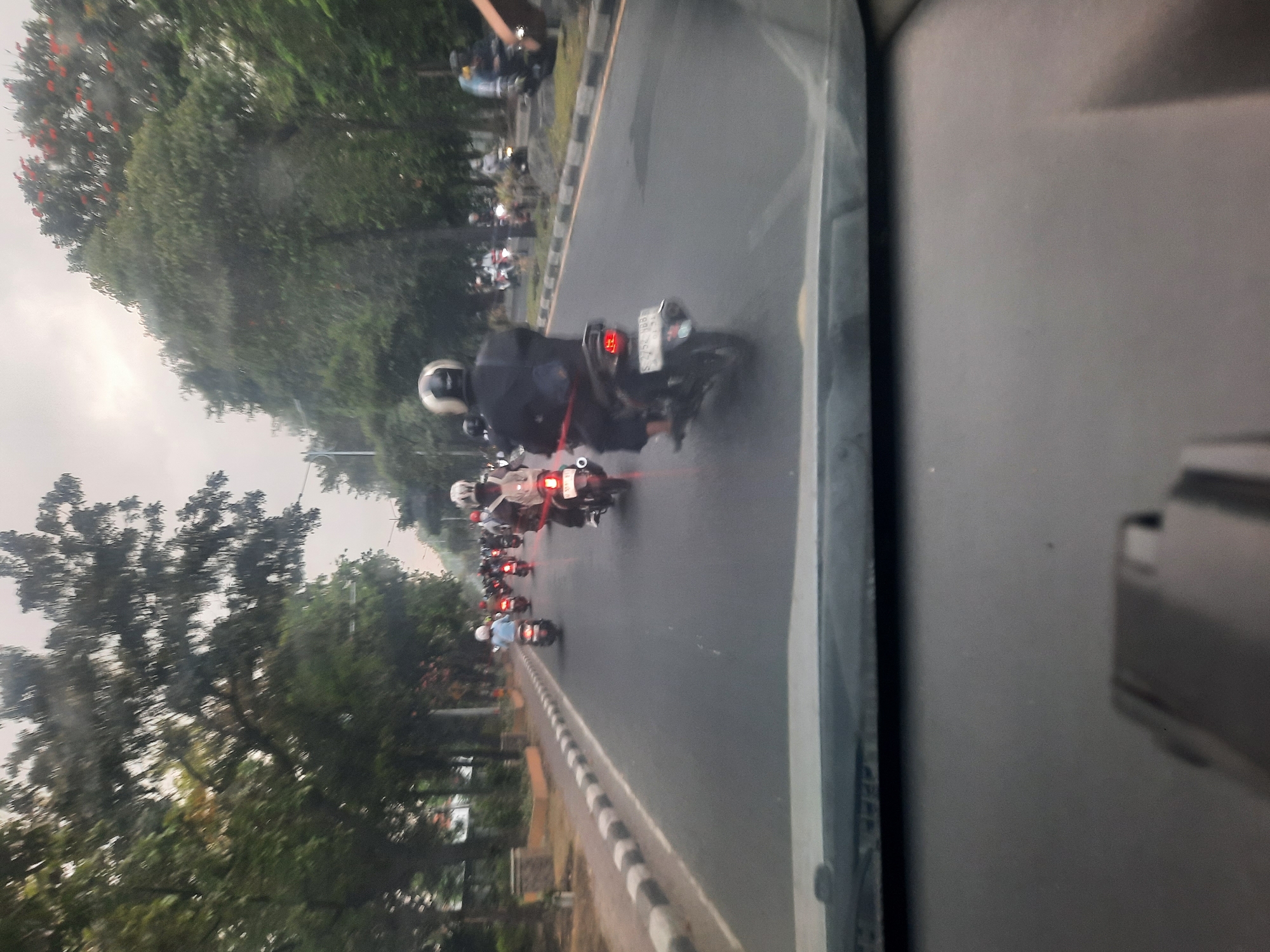 The image shows a street scene in Banjarkemantren, Indonesia, with numerous motorcycles on the road. A car is stationary in the foreground, with its windshield and dashboard visible, suggesting the photo was taken from inside a vehicle. The road is paved and lined with trees and some buildings on the left side. The sky is overcast, and the lighting is diffused. Several people are riding motorcycles, mostly wearing helmets, and some are carrying backpacks. The motorcycles are a mix of different models and colors, with their headlights and taillights illuminated. Some of the motorcycles have license plates visible. The overall impression is that of a busy street during the daytime, likely with moderate traffic.