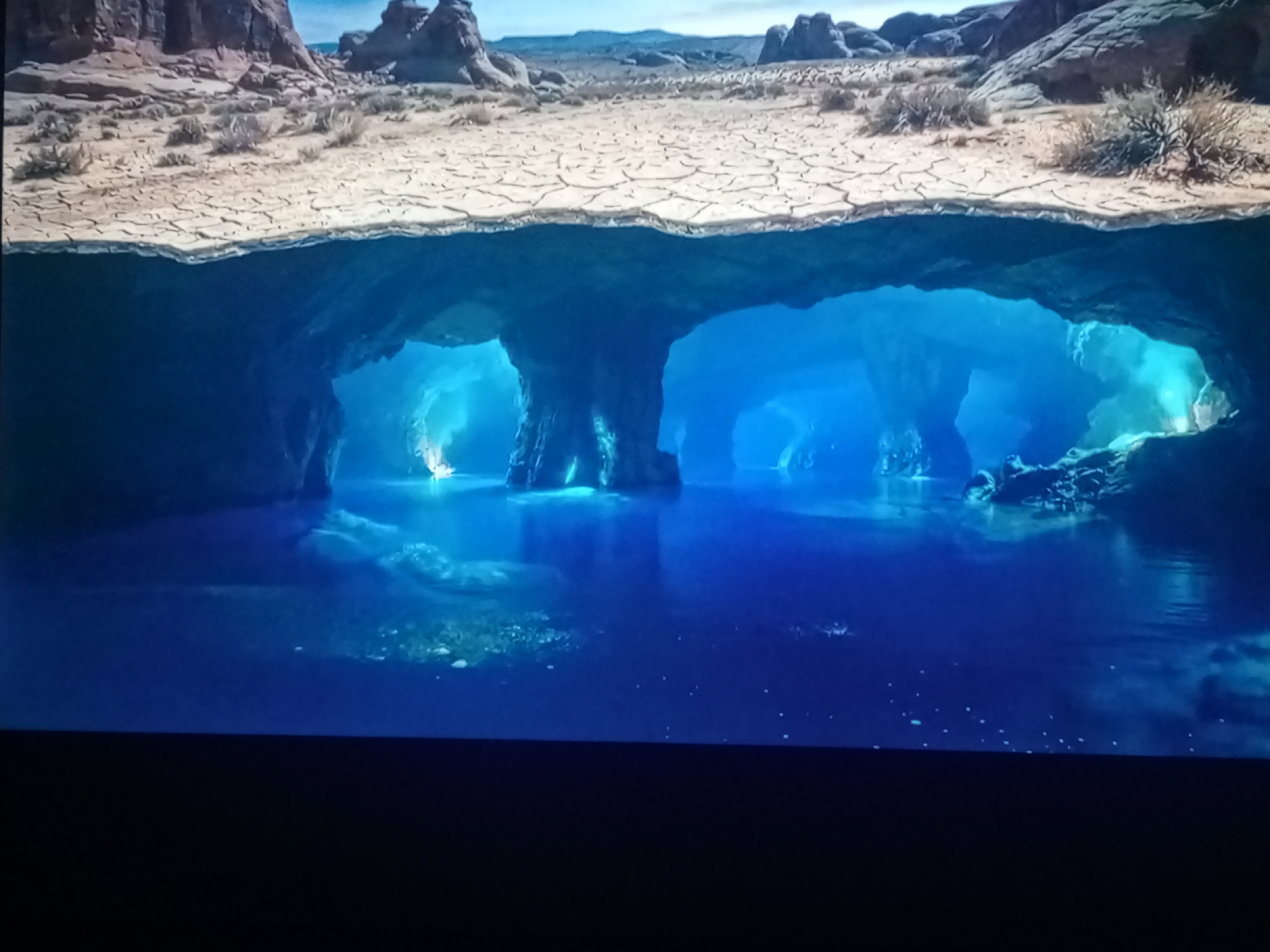 The image shows a split view of a dry, cracked desert landscape above and a submerged cave system with bright blue water below. The desert is arid with sparse, dry vegetation and rock formations in the background under a clear sky. The cave system features illuminated underwater caverns and rock pillars, with light filtering through the water, creating an ethereal blue glow. The water appears calm and clear, with some debris or sediment visible on the bottom in certain areas. There are no people or animals present in the scene. The scene is a surreal and fantastical depiction of an underground water system beneath a desert.