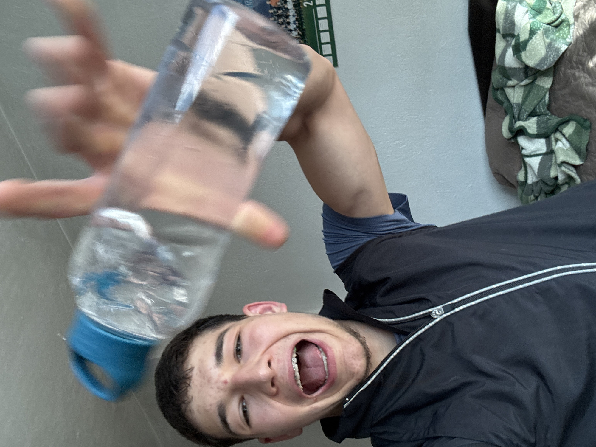 A young male is shown in a selfie-style image. He is holding a clear water bottle with a blue cap. His mouth is open, revealing braces. He is wearing a black jacket. The background is a light wall. There is a calendar hanging on the wall and a patterned blanket or cloth partially visible in the upper right corner. The location is not discernible as Zlatograd, Bulgaria.