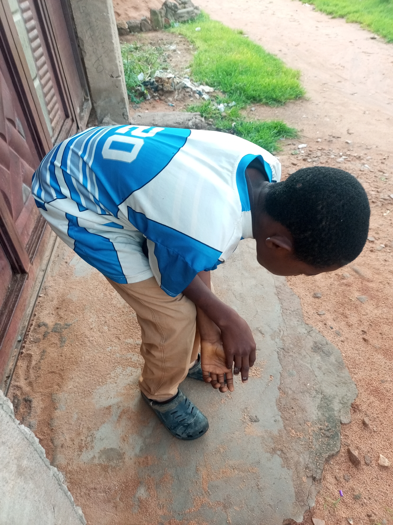 A young person wearing a white and blue jersey and tan pants is bending over, facing downwards. Their hands are clasped in front of their thighs. They are standing on a dirt and concrete surface next to a wooden structure and a patch of grass. The scene appears to be outdoors in what could be a residential area in Tempure, Nigeria, possibly on a path or near a building.