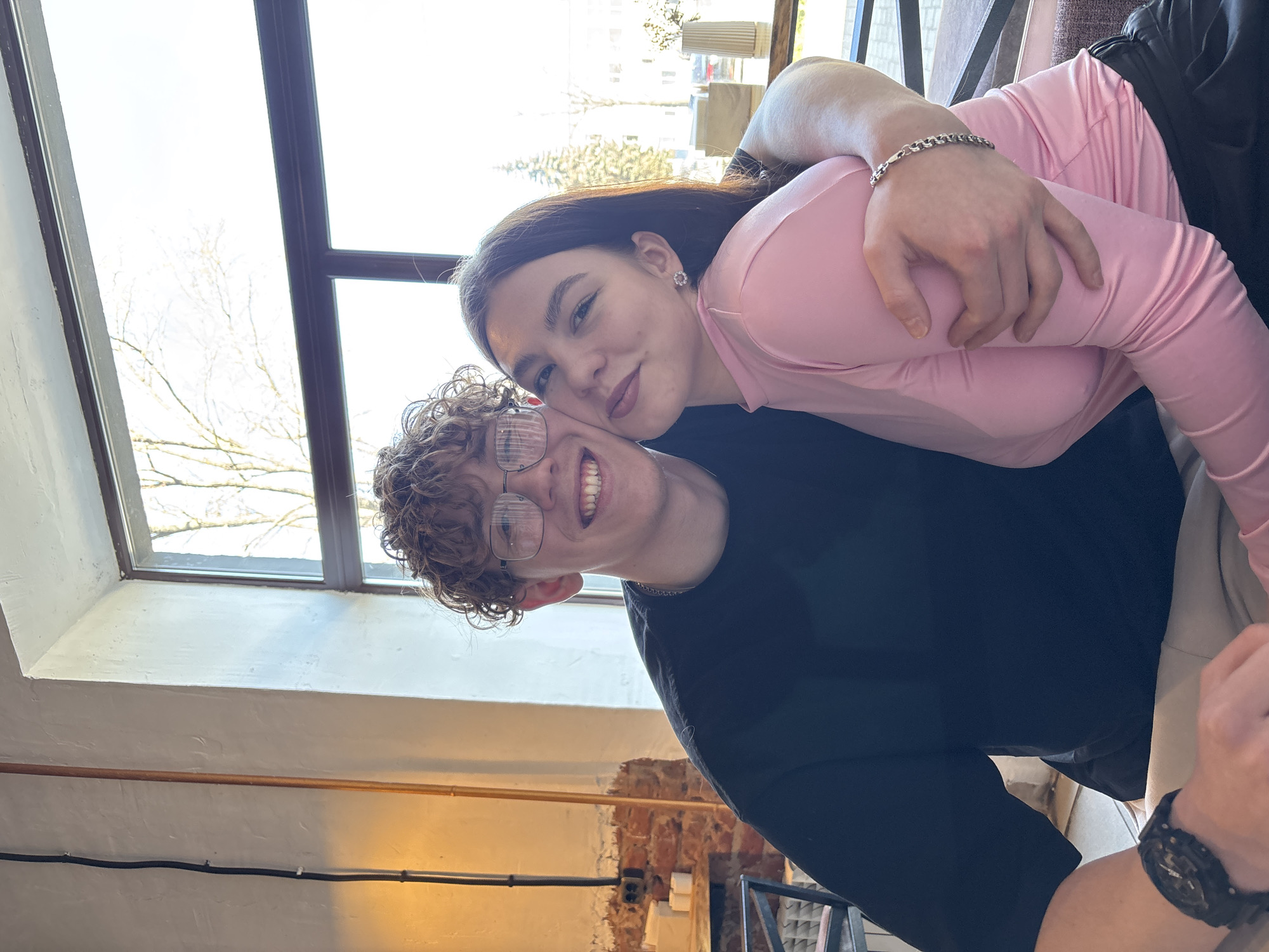 Two people, one wearing glasses and a black shirt and the other with dark hair and a pink shirt, are embracing. The location is Chaikovsky, Russia. A window is visible in the background, showing a building and bare trees. The wall and radiator are also visible in the background. The person in pink is wearing a silver bracelet. A black watch is visible on the wrist of someone sitting in the corner of the frame.