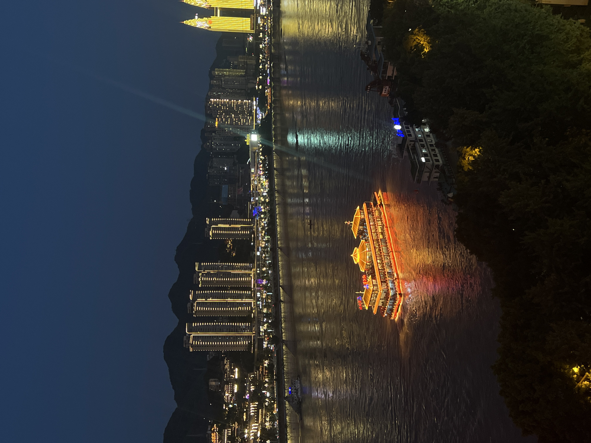 The image depicts a nighttime cityscape along a wide river, likely the Yangtze River in the Yuzhong District of Chongqing, China. The river occupies the majority of the frame, reflecting the lights of the city that line its banks.
Dominating the river's surface is a large, multi-decked passenger boat, illuminated with a vibrant red and yellow glow. The boat has a traditional architectural style with ornate roofs and balconies, suggesting it is a tour or dinner cruise vessel. It is moving, creating a visible wake.
On the far bank, a dense collection of modern high-rise buildings rises, outlined with bright, vertical lines of light. These buildings are characteristic of a bustling metropolis. Further to the right, two exceptionally tall, slender buildings are illuminated in a bright yellow, appearing to be architectural landmarks. A beam of light, possibly from a spotlight, emanates from a point within the cityscape and travels upwards into the dark sky.
The riverfront is lined with a promenade, also lit, with numerous smaller boats docked or moving. In the foreground, obscuring the lower right portion of the frame, is a dark, dense cluster of trees, possibly part of a park or hillside. Some scattered yellow lights are visible within the foliage, indicating further illumination or structures within the treeline. The sky is a deep, clear blue-black, indicative of nighttime. The overall scene conveys a sense of urban vibrancy and activity at night.