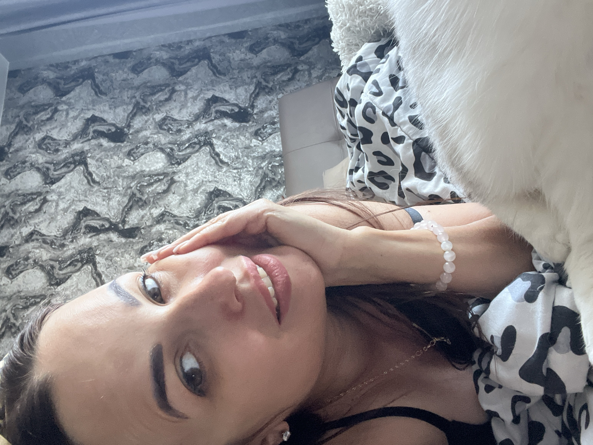 The image shows a close-up, low-angle selfie of a woman lying down, partially covered by a white, fluffy object, possibly a pet. She has long brown hair and is wearing a black top and a bracelet with light pink beads. Her right hand is covering part of her face, with her thumb touching her forehead and fingers resting on her cheekbone. She has brown eyes and is smiling, showing her teeth. The background appears to be a bedroom, with a textured wallpaper on the wall and a patterned duvet or blanket. The lighting suggests it is daytime. The overall mood is relaxed and intimate. There is no visible text in the image.