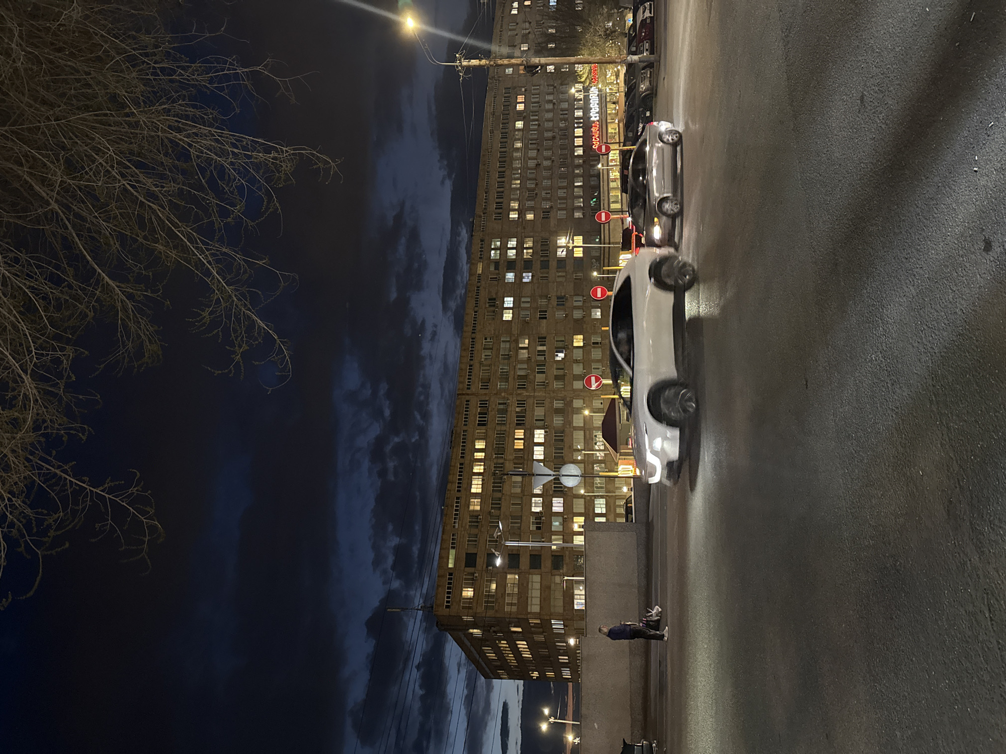 A long, tall building with a lit-up sign written in a foreign language dominates the background of a night scene. Numerous illuminated windows speckle the building. A stop sign is also visible. The right side of the image shows a portion of a street with two cars parked and a person standing on the sidewalk. The sky is dark with some clouds. A bare tree is on the left side of the image. The location is Yerevan, Armenia.