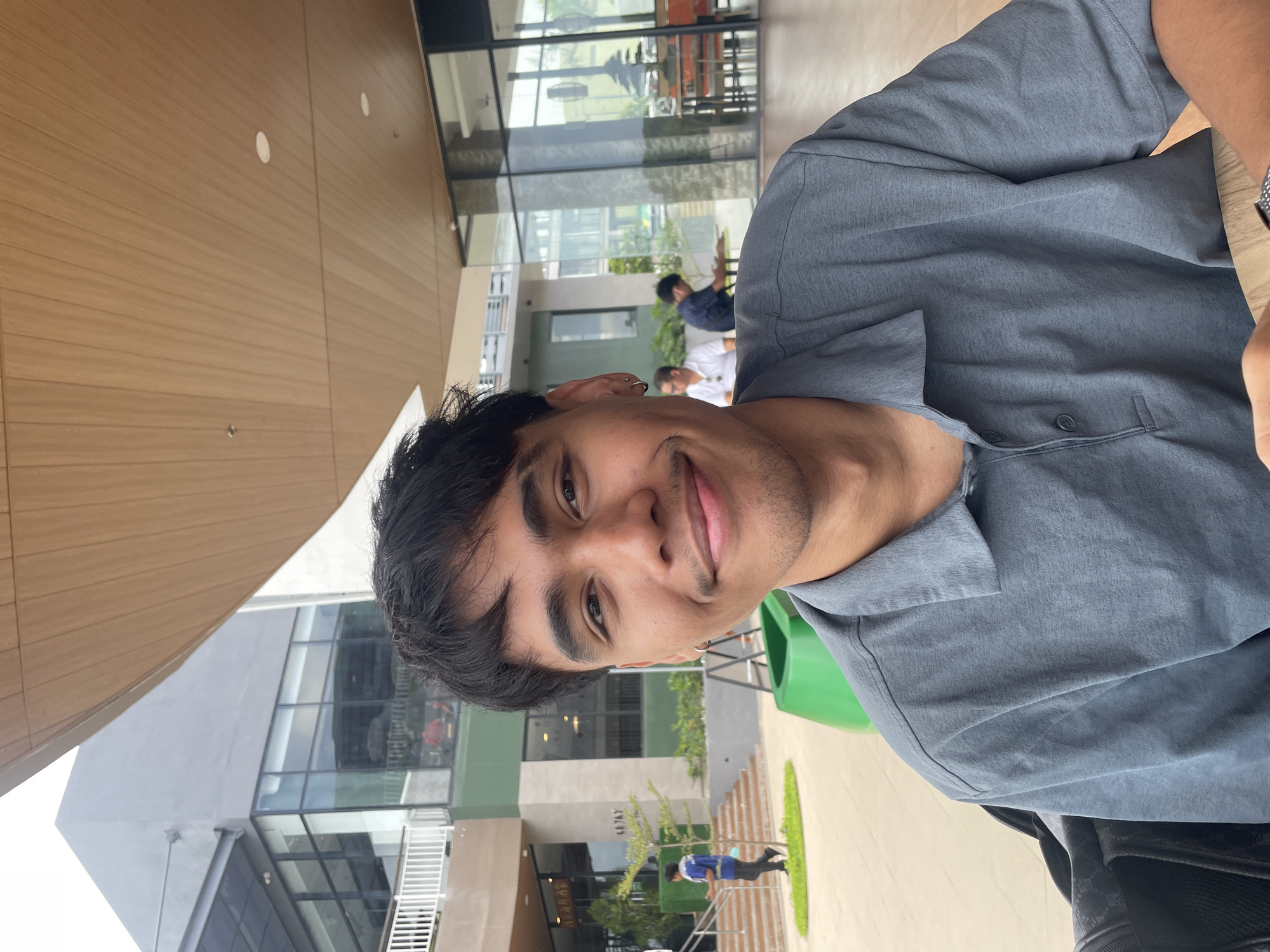 A young Filipino male is captured in a close-up selfie, seated outdoors. He has dark, short, messy hair, brown eyes, and a faint mustache. He is wearing a grey collared polo shirt. Behind him, a modern building with large glass windows and a wooden-slatted overhang is visible. In the background, two other individuals are seated at a table, and another person is walking up a set of stairs. A bright green planter is also in the foreground. The location appears to be an outdoor seating area of a commercial establishment in Davao City, Philippines, possibly within a mall or a similar urban development.