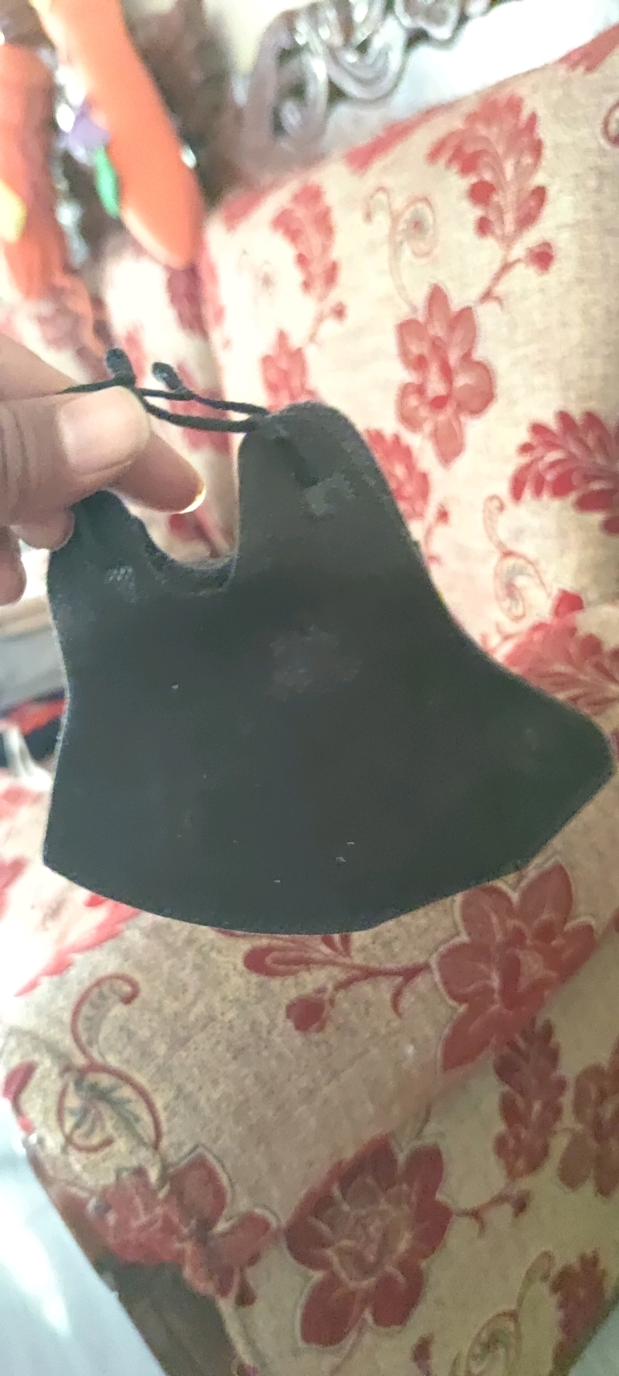 A close-up shot shows a black fabric object, possibly a mask or a piece of clothing, held by a person's hand. The object has a distinctive shape with curved edges and appears to be made of a soft material. Two thin black strings are attached to the top of the object, held by the person's thumb and index finger. The background is blurred but reveals a patterned fabric, likely a sofa or cushion, with floral designs in shades of red and cream. The lighting suggests an indoor environment. The image does not provide any information about the city of Gambarsari, Indonesia.