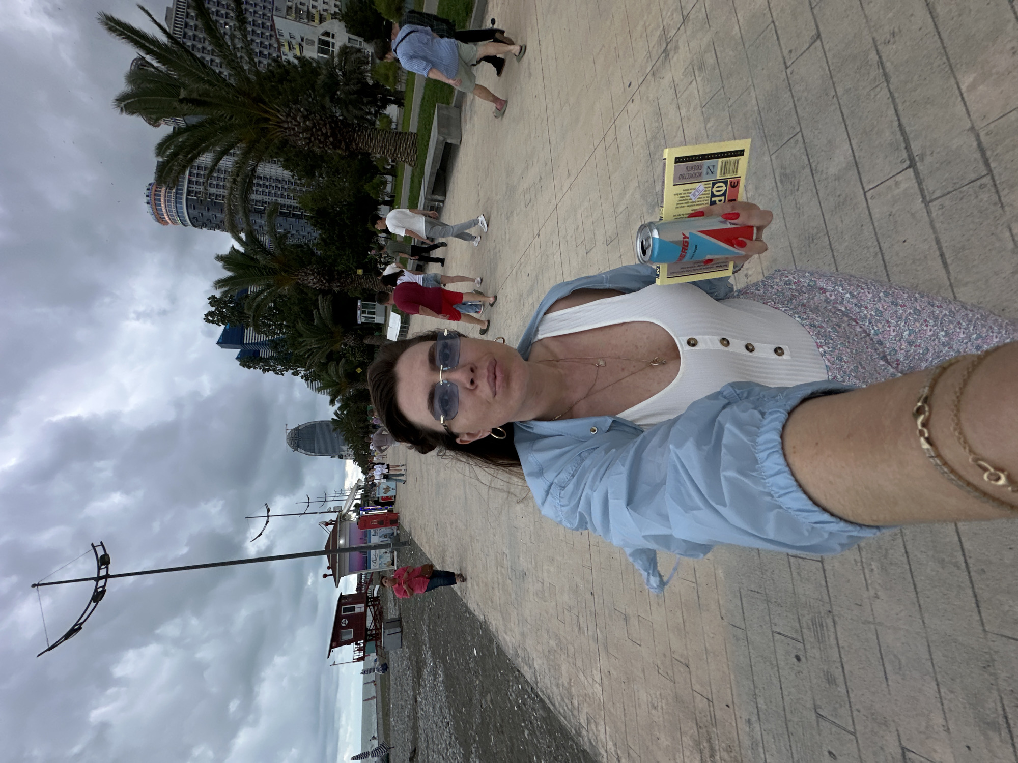 In Batumi, Georgia, a woman with sunglasses and a light blue jacket takes a selfie on a paved walkway adjacent to a beach. She holds a can of "Energy Drink" and a yellow pamphlet. Several people are visible in the background walking along the walkway and on the beach, with palm trees and modern buildings lining the street behind them. The sky is overcast, indicating cloudy weather.