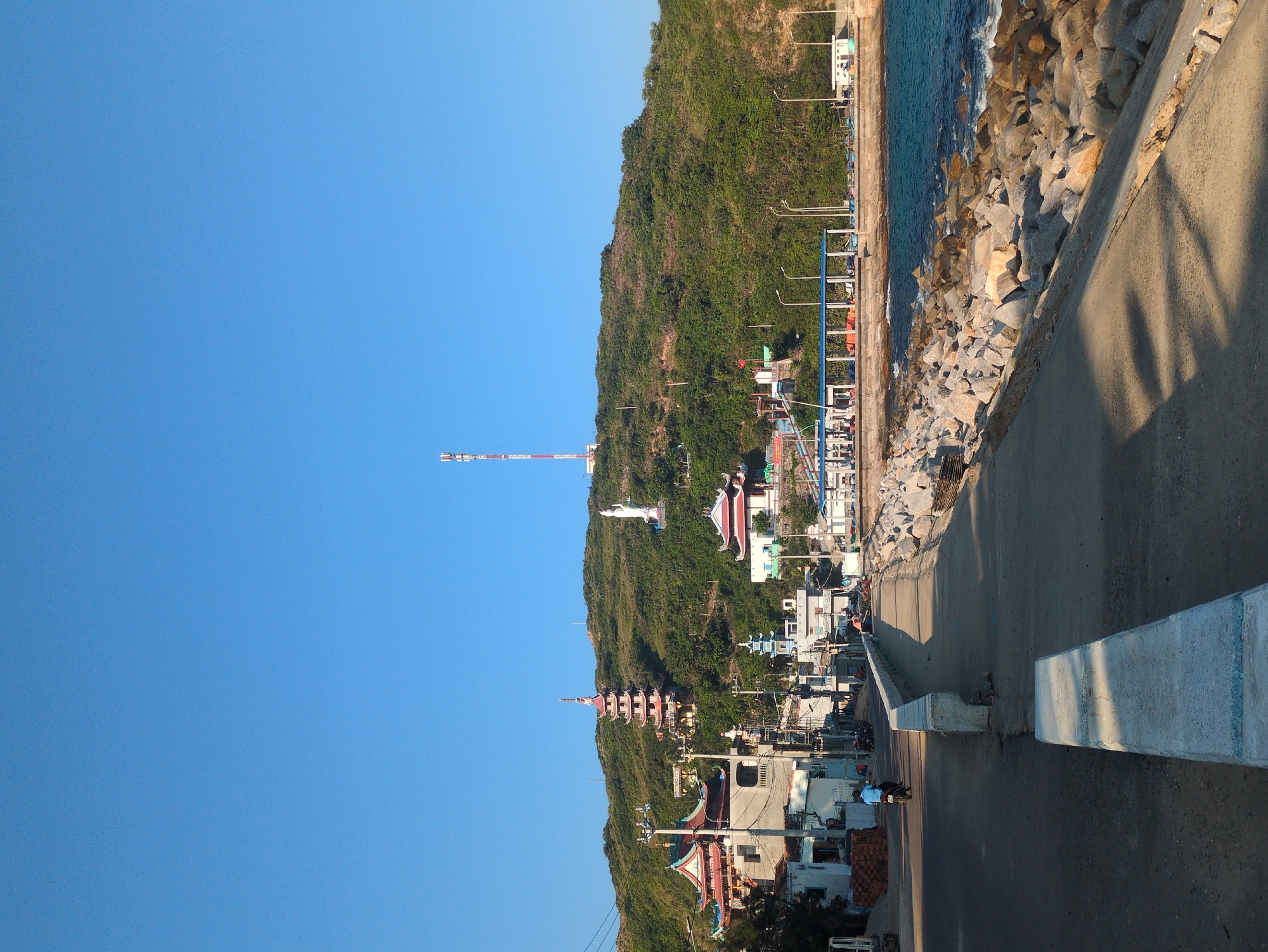 The image depicts a coastal urban-rural transition zone in East Quy Nhon Ward, Vietnam, under a clear blue sky. In the foreground, a paved road with a raised concrete median extends from the lower-left, curving gently to the mid-right. To the right of the road, a concrete seawall protects a rocky breakwater where turquoise-blue water, agitated by small waves, meets the land.
In the midground, a lone individual on a dark motorcycle, wearing a light blue shirt, travels away from the viewer on the paved road. Along the left side of the road, a series of light-colored, multi-story buildings, some with traditional Vietnamese architectural elements and red roofs, line the street. Stacks of bricks are visible on the roadside near these buildings, indicating construction or storage. Further along the coastline, to the right of the road, a blue-roofed, open-sided structure, possibly a market or shelter, is situated near the water, with several small fishing boats visible nearby.
The background is dominated by a steep, heavily vegetated green hill. Prominently situated on the lower-mid slope of the hill is a multi-tiered pagoda featuring red and gold accents. Higher and to the right of the pagoda, a large white statue of a deity (likely Guan Yin/Avalokiteshvara) stands. At the summit of the hill, a tall, red and white striped cellular telecommunications tower is clearly visible against the sky. Utility poles with numerous wires are distributed throughout the scene on the left, and streetlights are visible near the coastal structures. The scene is brightly lit by direct sunlight, casting shadows to the right, suggesting morning or early afternoon.