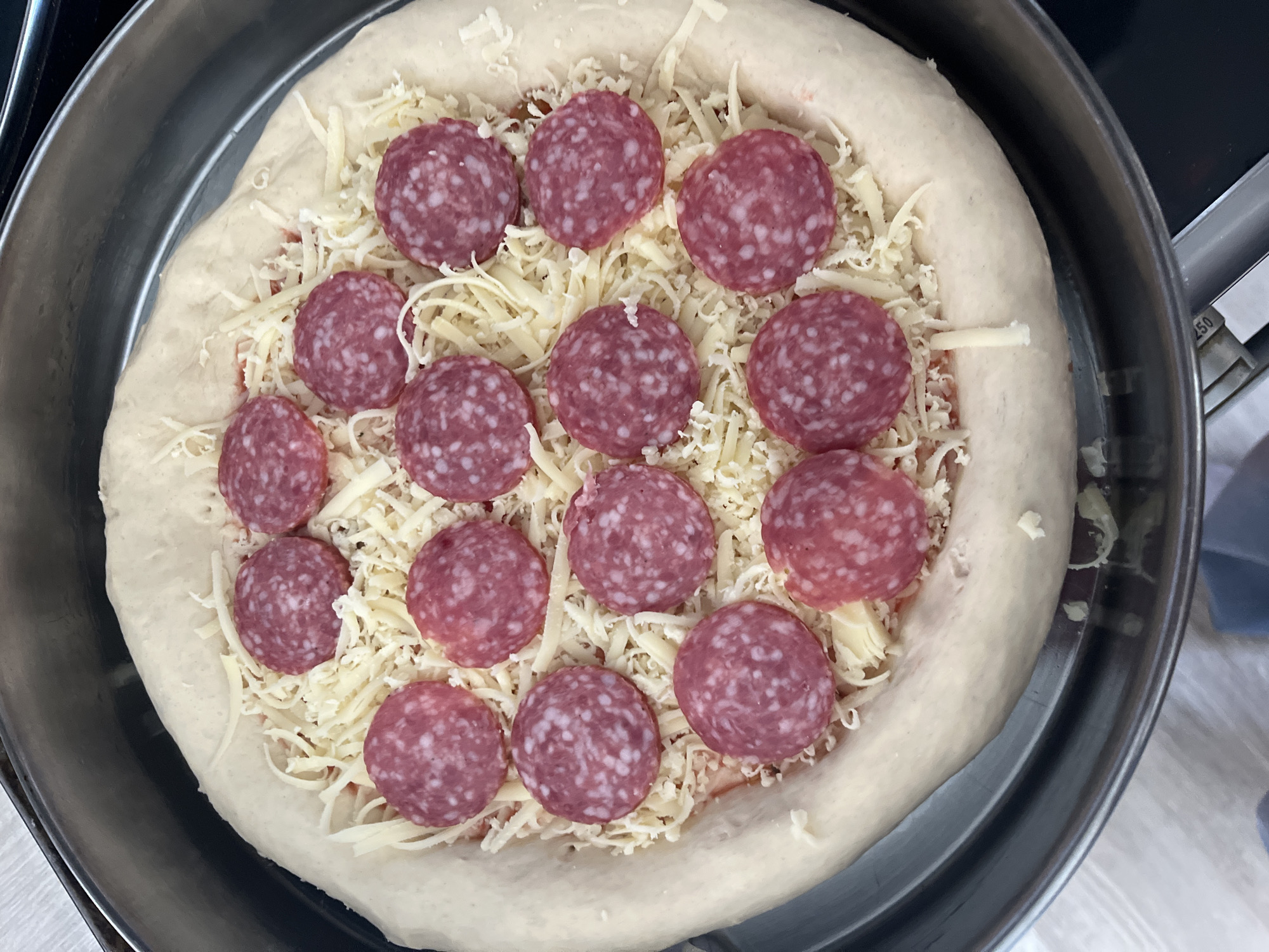 A top-down, close-up shot shows an uncooked pizza in a metal pan. The pizza has a thick, raised crust. The pizza is topped with tomato sauce, shredded white cheese, and 13 slices of pepperoni arranged in a circular pattern. The pan is sitting on a dark-colored surface, possibly a stovetop. The pizza is centrally located in the image, filling most of the frame. The specific location within Burgas, Bulgaria, cannot be determined from the image.