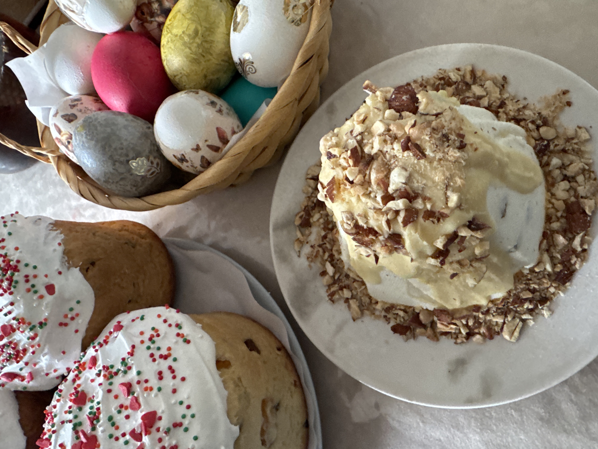 An eye-level shot depicts an Easter feast featuring a basket of decorated eggs, kulich cakes, and paska on a light-colored tablecloth. A woven basket holds a variety of painted eggs, including white, pink, green, gray, and blue hues, some adorned with patterns. To the left are kulich cakes, covered in white icing and colorful sprinkles. On a white plate is paska, topped with chopped nuts and creamy frosting. The scene is likely set indoors during Easter celebrations. Chaikovsky, Russia, is not discernible from the image.