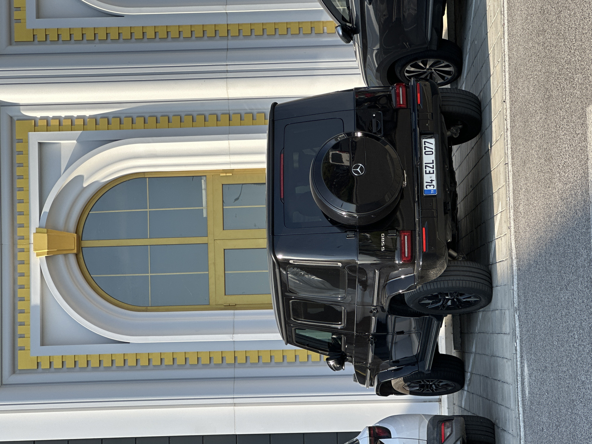 The image captures the rear three-quarter view of a black Mercedes-Benz G-Wagen, model G 580, parked on light grey pavers in front of a distinct architectural building. The vehicle displays a Turkish license plate, "34 EZL 077", indicating its registration in Istanbul, Turkey. A black spare tire cover, bearing the Mercedes-Benz logo, is mounted on the rear door. Black multi-spoke alloy wheels are visible on both the front and rear axles. To the left, a portion of a white SUV is visible, showing its rear quarter panel. To the right, part of a dark-colored SUV is partially visible, showing its front wheel and side.
The background features a building with a light grey or off-white facade, accentuated by prominent gold/yellow decorative elements. A large, arched window or doorway with a golden frame and internal mullions is centered behind the G-Wagen. The building's architecture includes vertical fluted pilasters and decorative rectangular gold blocks arranged in a repetitive, stacked pattern along vertical edges. Arched architectural details above the windows/doorways also feature golden accents. The scene is illuminated by strong, natural sunlight, casting defined shadows. In the immediate foreground, an asphalt road surface is visible, separated from the paved parking area by a thin white line. No individuals are present in the image. The specified location of Kapitan Andreevo, Bulgaria, is consistent with the presence of a Turkish-registered vehicle, given the town's position at the Bulgarian-Turkish border.