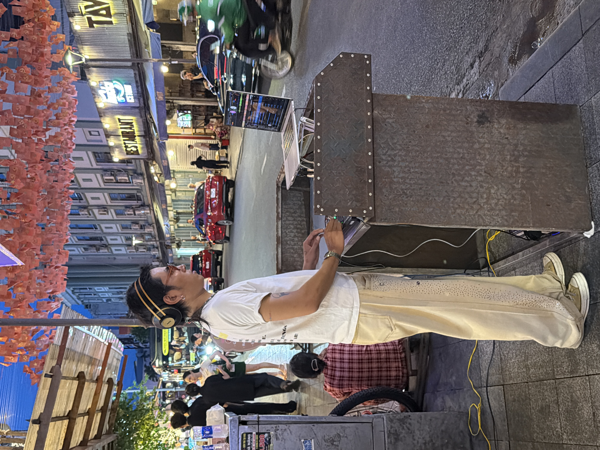 In Thuận Hóa Ward, Vietnam, a DJ, wearing headphones, a white t-shirt, and tan pants adorned with rhinestones, operates a laptop connected to DJ equipment. The DJ stands on a sidewalk next to a rough, metal DJ booth. Behind the DJ, a person wearing a red and white plaid shirt sits on a low stool. The street in front of the sidewalk features several vehicles, including red cars and a motorcycle with a rider in a green uniform, suggesting a bustling urban environment. Buildings with illuminated signs line the street, and festive orange paper decorations hang overhead, indicating a lively, possibly commercial or entertainment district.