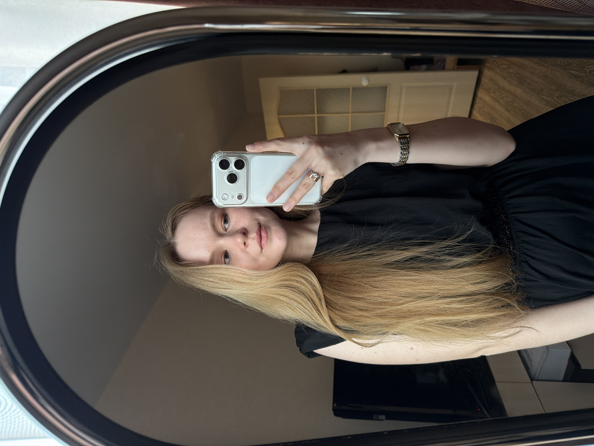 A woman is taking a selfie in a mirror. She has long, blond hair and is wearing a black dress and a watch. She is holding a white smartphone. The background appears to be a room with a door. The location in the city of Izhevsk, Russia, cannot be determined from the image.