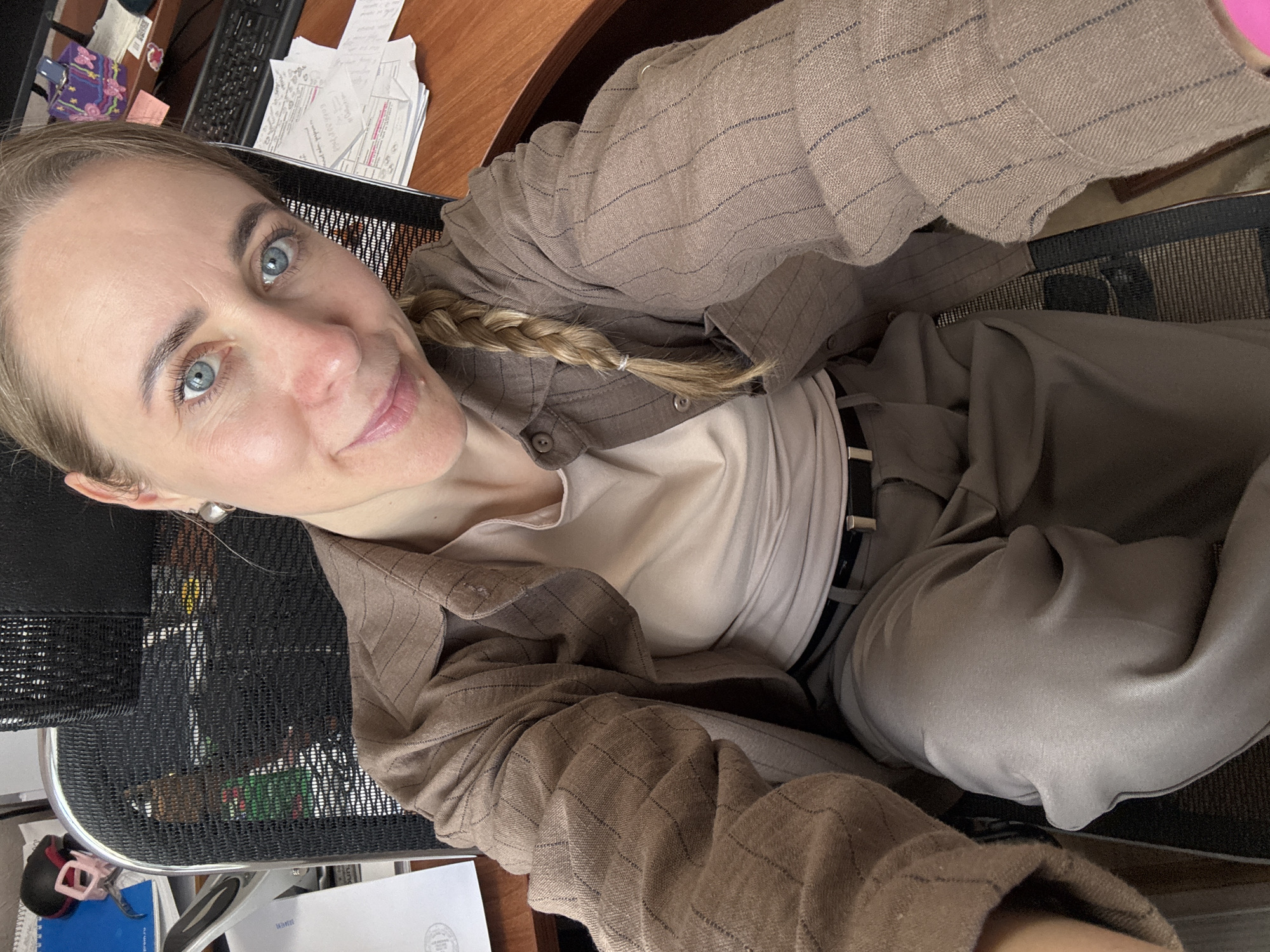 A woman with light skin and blue eyes sits in a black mesh office chair. Her blonde hair is pulled back in a braid. She wears a light-colored shirt, a brown pinstripe jacket, and matching pants, secured with a black belt. She appears to be seated at a desk with papers and office supplies. The location is likely an office or workspace, but there is no visual information to confirm it is in Chaikovsky, Russia.