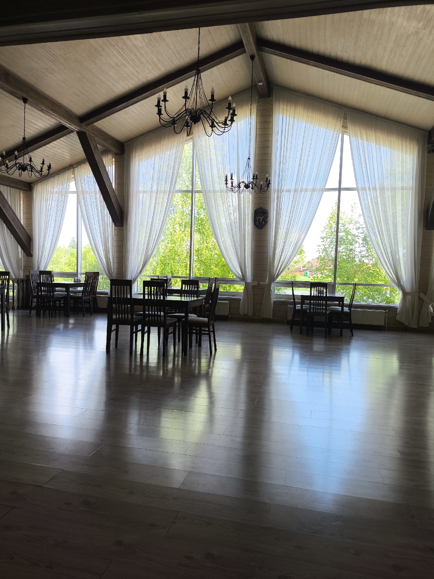 The image shows a spacious, well-lit interior with a high, sloped ceiling supported by exposed wooden beams. Two elaborate black chandeliers hang from the ceiling. Large, floor-to-ceiling windows, draped with sheer white curtains, dominate the far wall, offering a view of lush green trees and a bright sky. The room is furnished with several dark wooden tables, each surrounded by matching chairs, arranged as dining or seating areas. The floor is a polished, reflective dark wood. The overall impression is that of a restaurant, banquet hall, or a similar venue designed for gatherings. The location is specified as being in the city of Taldom, Russia.