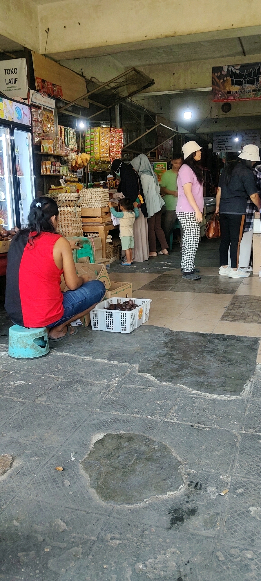 The image depicts an outdoor market scene in Mekarmukti, Indonesia, likely a section of a busy street or an open-air market stall. In the foreground, a person wearing a red tank top and blue jeans squats on a small blue stool next to a white plastic crate filled with dark, oval-shaped produce, possibly dates or plums. The ground is paved with textured, square tiles, some of which are discolored and wet, indicating a recently cleaned or leaky area.
Behind this individual, a market stall is visible, displaying various goods. Stacked cardboard boxes are filled with rows of eggs, and shelves are stocked with packaged food items in colorful wrappers. Several people are browsing or working at the stall. A woman wearing a light-colored headscarf and a light blue hijab stands near the egg display, and a child in a light-colored shirt and shorts is reaching towards a shelf. Another person, also wearing a headscarf and black attire, is further back. To the right, two women in casual attire, one in a pink t-shirt and patterned pants, and another in a dark top and black pants, are facing away from the camera, seemingly engaging with the market offerings. A man in a green shirt is partially visible in the background. The market environment appears somewhat cluttered, with overhead structures and signage, including a sign that reads "TOKO LATIF." The overall impression is one of a typical, active local market.