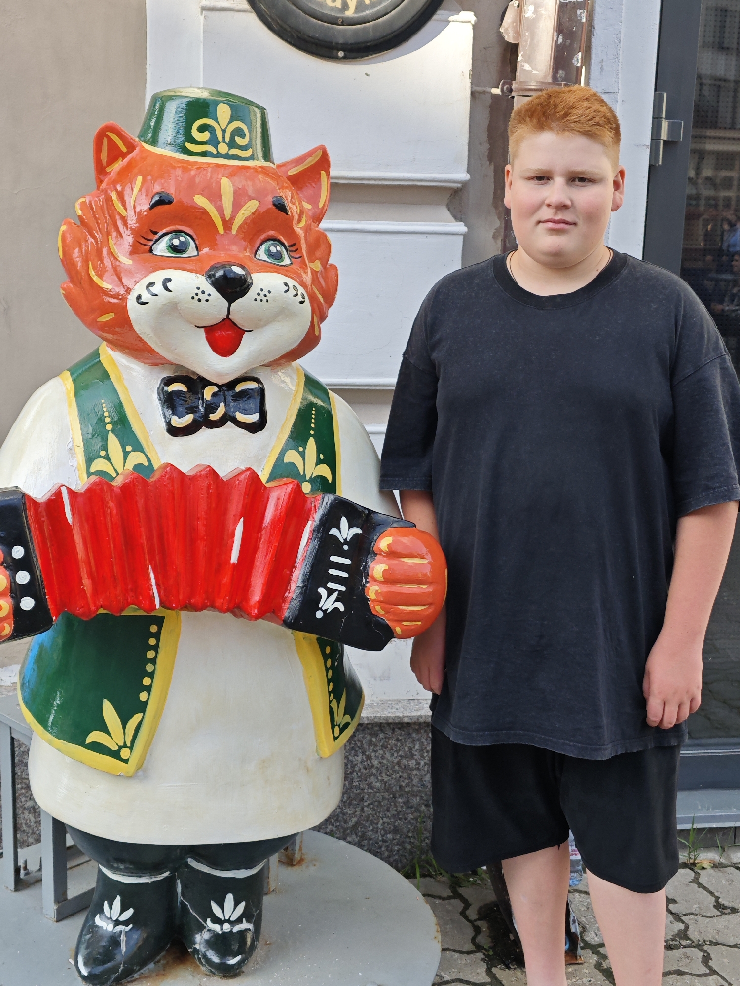 A young person with reddish-blond hair stands next to a colorful statue of a cat playing an accordion. The statue is vibrantly painted in orange, green, and yellow, with a black bow tie and boots. It wears a green hat with a yellow emblem. The cat statue is positioned on the left side of the image. The person, on the right, is wearing a dark gray t-shirt and black shorts. They have a neutral expression. The background shows a building with white architectural details and a gray wall. The ground is paved with flagstones. The scene appears to be outdoors, possibly on a street or in front of a shop, given the building facade and paved ground. The lighting suggests it is daytime. There is no visible text in the image. The context provided is Kazan, Russia, implying this may be a local landmark or art installation.