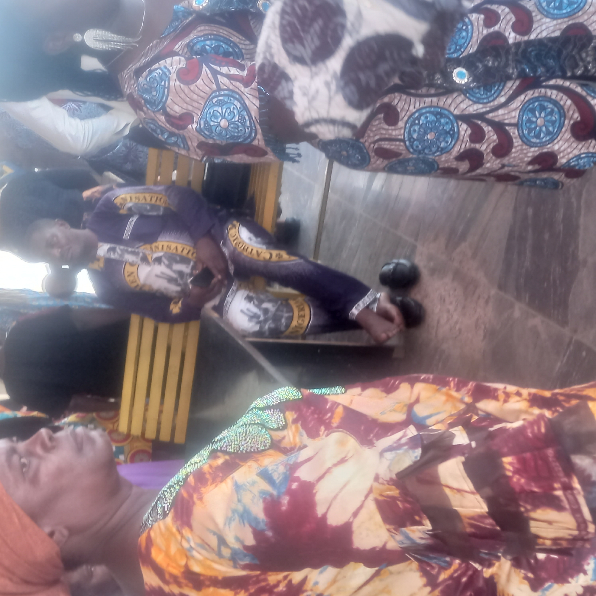 A group of people are gathered, possibly at an event. One person in a purple outfit lies on a yellow slatted bench. Another person wearing a patterned dress and head covering is visible at the bottom of the frame, also appearing to lie down. The location is likely Barden Yana, Nigeria, based on the prompt. Other patterned clothing is visible in the image.