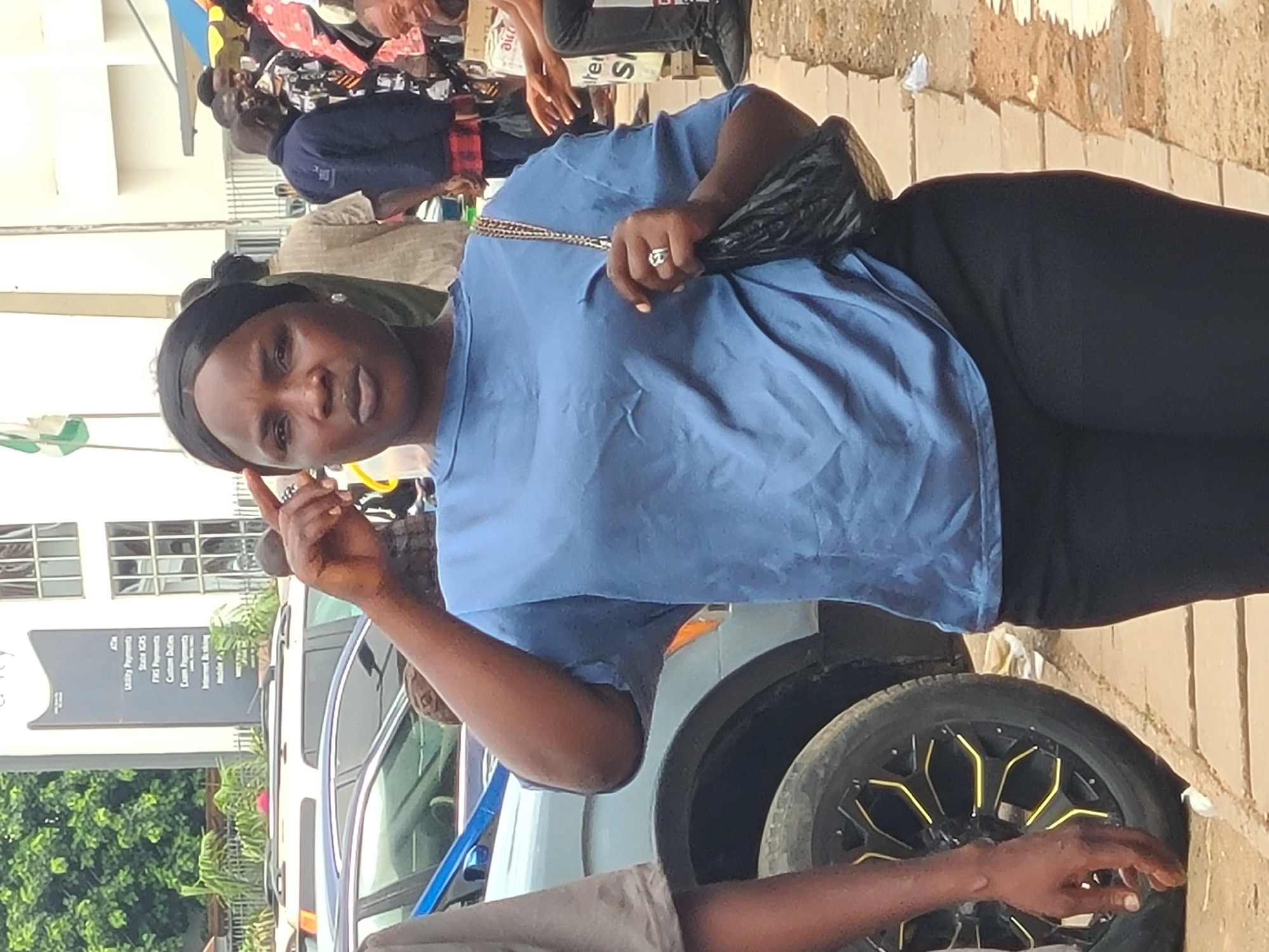 The image captures an outdoor scene, likely in a bustling urban environment, possibly Jos, Nigeria. A woman in the foreground is the central subject, dressed in a blue t-shirt and black trousers, with a black headscarf. She has a ring on her left hand and is holding a black bag. Her left hand is raised to her forehead, and her expression is contemplative. Behind her, several people are visible, suggesting a public space. Cars are parked in the background, and a building with signage is partially visible. The ground appears to be paved with concrete and dirt.