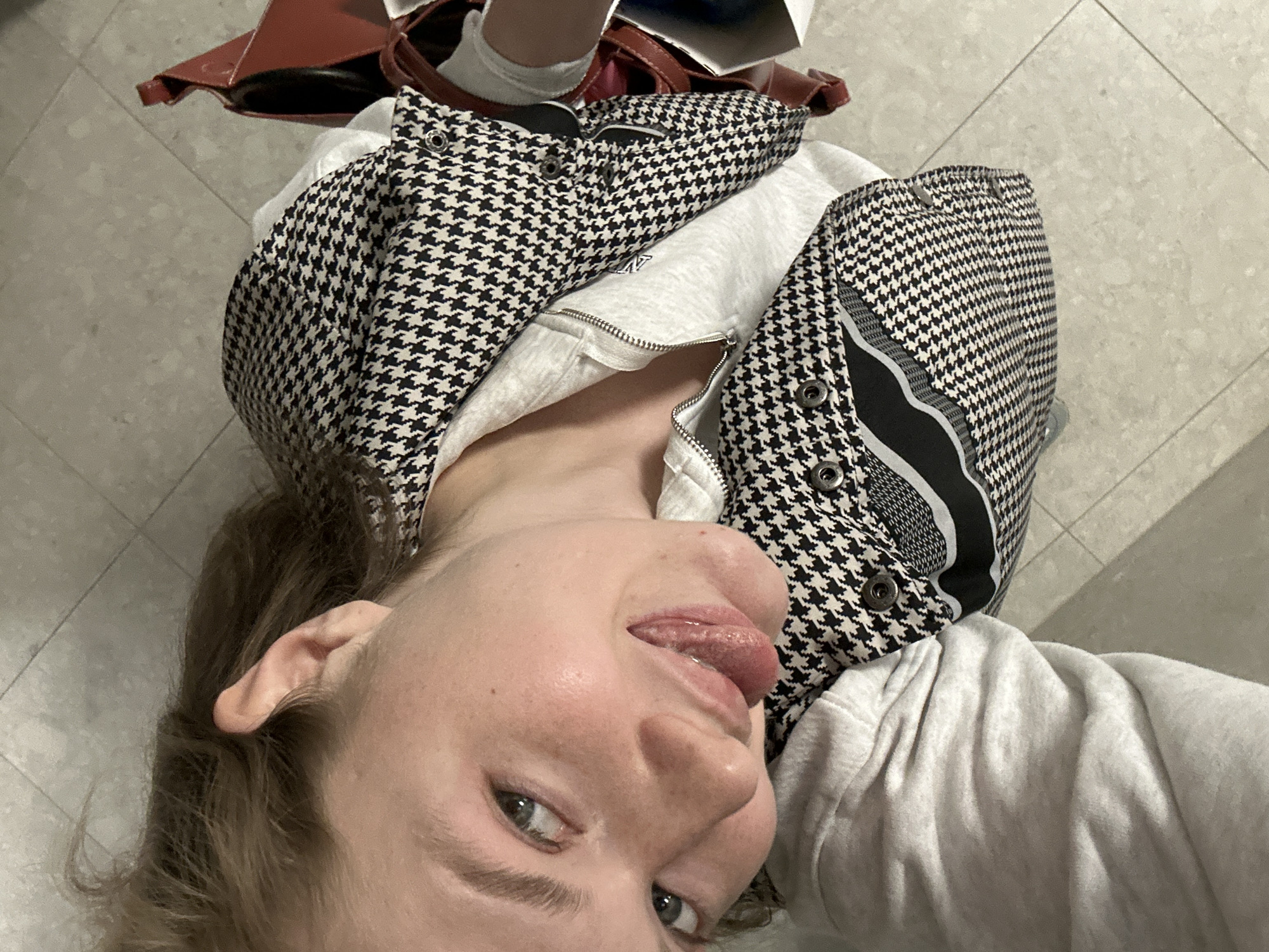 This high-angle selfie features a young person, likely female, situated indoors in Kazan, Russia. The individual is looking directly up at the camera, possibly while lying down or leaning back, and is making a playful expression with their tongue slightly extended.
The person has light brown hair and blue-green eyes. They are wearing a black and white houndstooth pattern jacket or hoodie, which includes metal snap fasteners on the front panels and a zipper. A light grey hooded sweatshirt is visible underneath or as part of the main garment.
In the upper background, a dark red or brown bag or purse is partially visible, along with what appears to be white socks. The setting features a floor composed of light-colored, square tiles. The overall mood conveyed by the subject's expression is casual and lighthearted. No discernible text is visible within the image.