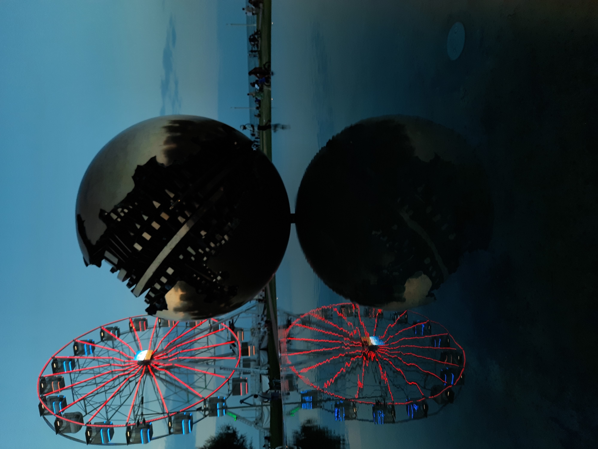 The image captures a twilight scene at the Piazza del Popolo in Pesaro, Italy. A large, spherical sculpture, possibly a representation of the Earth, dominates the foreground, its metallic surface reflecting the surrounding environment and the dusky sky. To the left, a Ferris wheel is illuminated with red and blue lights, its vibrant glow contrasting with the darkening sky. The reflections of both the sculpture and the Ferris wheel are clearly visible in a calm body of water in front of them. In the background, a few silhouetted figures can be seen gathered along a waterfront, possibly enjoying the evening ambiance. The overall atmosphere is one of serene leisure, with the artificial lights of the amusement ride and the sculpture creating a dynamic visual focal point against the natural backdrop of the sea and sky.