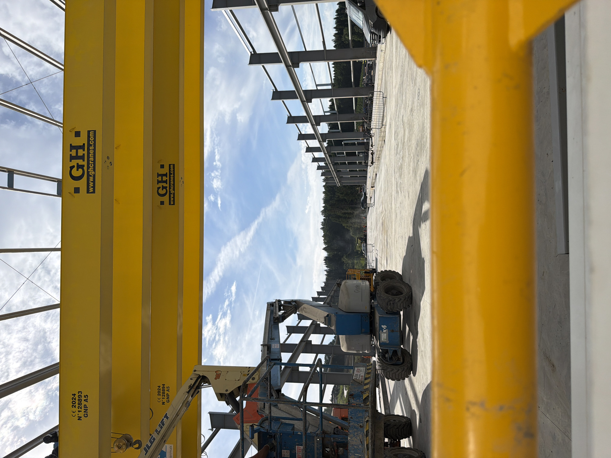 In Janowice, Poland, a construction site is captured under a partly cloudy sky. The foreground features a bright yellow overhead crane beam, obscuring the lower portion of the image. Two similar yellow beams run horizontally across the midground, each bearing the "GH" logo and the website "www.ghcranes.com". Below the top beam, the text "CE 2024", "N° 128893", and "GNP A5" is visible. The lower yellow beam also displays "CE 2024", "N° 128894", and "GNP A5".
To the left, a blue aerial work platform with the "JLG LIFT" logo is positioned. Next to it, another blue aerial work platform, this one a Genie brand, is visible with "x4" marked on its side. In the background, the skeletal structure of a large building is under construction, with numerous steel beams erected. A dark-colored van is parked on the right side of the construction area. The ground is paved, and a line of trees forms the treeline in the distance.