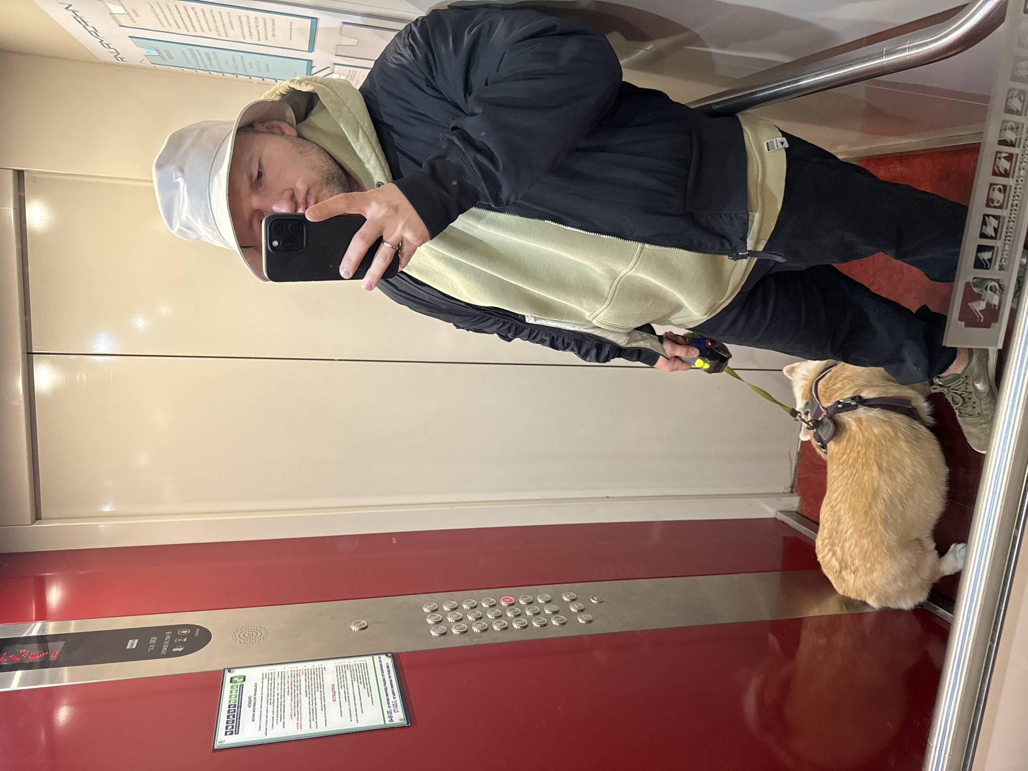 A person wearing a white bucket hat, a green hoodie, and a black jacket holds a phone and stands in an elevator. A corgi dog is on a leash next to the person. The elevator has red and white walls, a metal rail, and a panel of buttons. The elevator appears to be located in Krasnoyarsk, Russia.