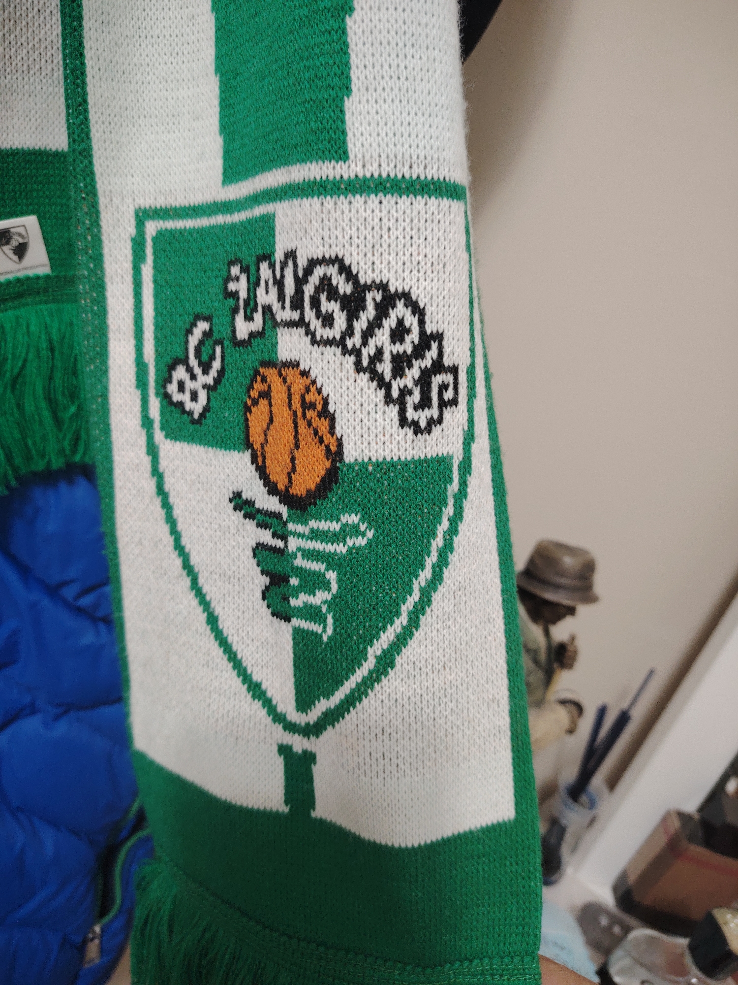 The image displays a close-up of a knitted sports scarf, primarily in green and white. The scarf features a prominent shield-shaped emblem that is vertically divided into green and white sections, outlined in contrasting colors. At the top of the emblem, the text "BC ŽALGIRIS" is knitted in black and white. Below the text, an orange basketball with black outlines is depicted. The lower section of the emblem displays a stylized green symbol resembling a plant or branch against a white background. The scarf has green fringes visible on its left side.
To the immediate left of the scarf, a blue quilted or puffer garment is partially visible and out of focus. The background to the right of the scarf consists of a plain, light-colored wall. Further in the background, out of focus, a small figurine or statue wearing a brown hat and light-colored clothing is discernible, along with various indistinct objects on a surface, including what appear to be pens or brushes in a container.
Regarding the location in Huntingdonshire, United Kingdom, there is no visual information within the image to suggest or confirm this specific geographical location. The scarf explicitly displays the name "BC ŽALGIRIS," which is a well-known Lithuanian professional basketball club, indicating a potential connection to Lithuania rather than Huntingdonshire.