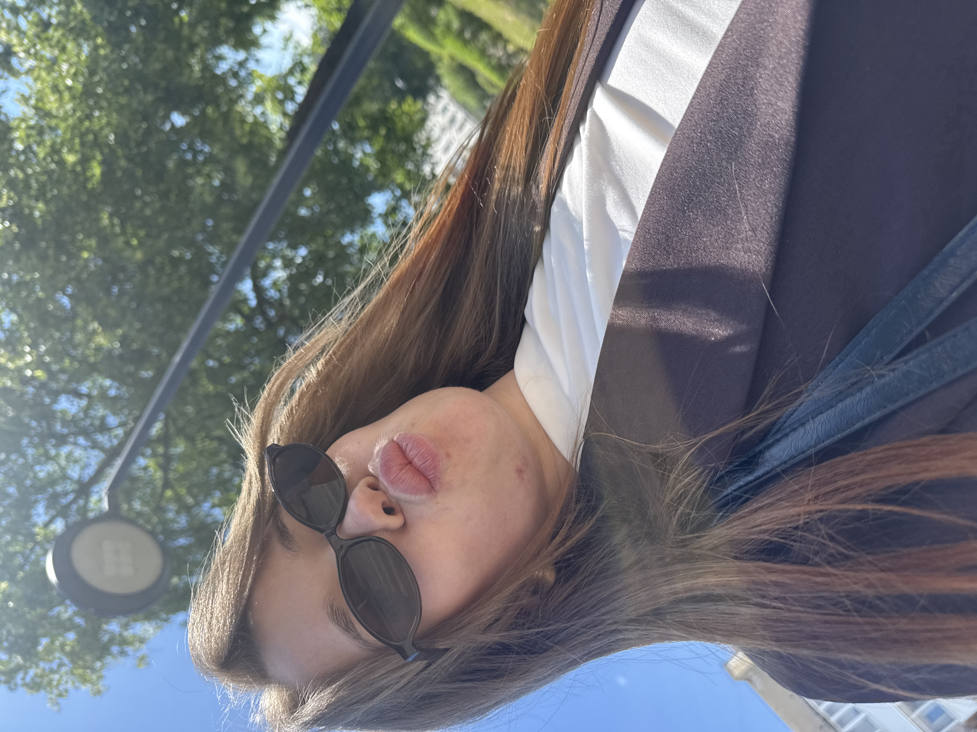 A person is captured in a low-angle close-up, outdoors in the city of Chaikovsky, Russia, under clear blue skies and bright sunlight. The individual, primarily featuring from the forehead down to the mid-torso, is wearing dark, oval-shaped sunglasses that obscure their eyes and upper nose. Their lips are pursed in a distinct "kissing" or "pouting" expression. Light brown hair with warmer highlights cascades down, framing the face and resting on a white garment. A dark brown or deep plum blazer or jacket is worn over the white garment, with a dark strap, possibly from a bag, visible across the jacket on the left side. The bright sunlight creates visible highlights on the hair and fabric, and there is a lens flare in the lower right quadrant. In the blurred background, a street lamp with a circular fixture is prominent in the upper center, positioned against a clear blue sky. Lush green trees and foliage are visible in the mid-background, out of focus. A light-colored building with multiple windows is partially visible on the far left, adding to the urban context.