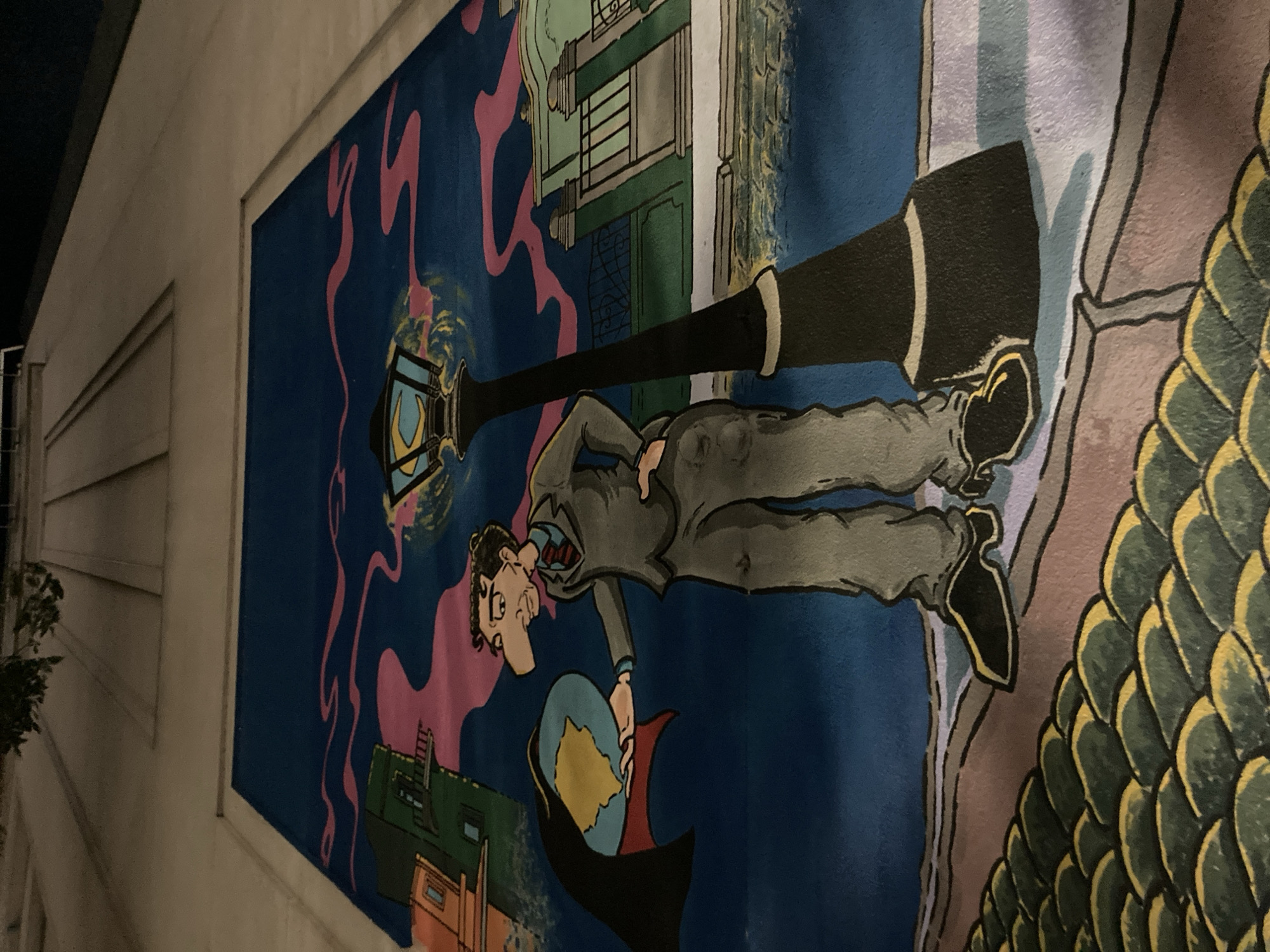 The image displays a vibrant mural painted on an exterior wall, viewed at what appears to be night, indicated by the dark surroundings outside the mural and the lit lamppost within the artwork. The mural itself is oriented vertically, featuring a central male figure in a cartoonish, illustrative style.
The man, dressed in a grey suit, white shirt, and red tie, has dark hair, glasses, and a distinctive long nose. He is depicted holding a small globe of the Earth in his left hand, gazing down at it with a contemplative or concerned expression.
Behind and slightly above the man is a tall, black lamppost with a glowing, ornate lantern, casting a blueish light. The background of the mural is a deep blue, adorned with dynamic, swirling pink patterns that could suggest smoke, clouds, or mystical energy. Below these patterns, stylized buildings with green rooftops and windows are visible, adding to the urban or fantastical setting.
On the right side of the mural, a large, intricate pattern of overlapping green and yellow scales dominates the foreground, resembling the skin of a dragon or a large mythical fish. The mural is bordered by a light-colored building façade, with horizontal siding visible on the left side of the frame, contrasting with the dark night sky above. There is no visible text within the mural.